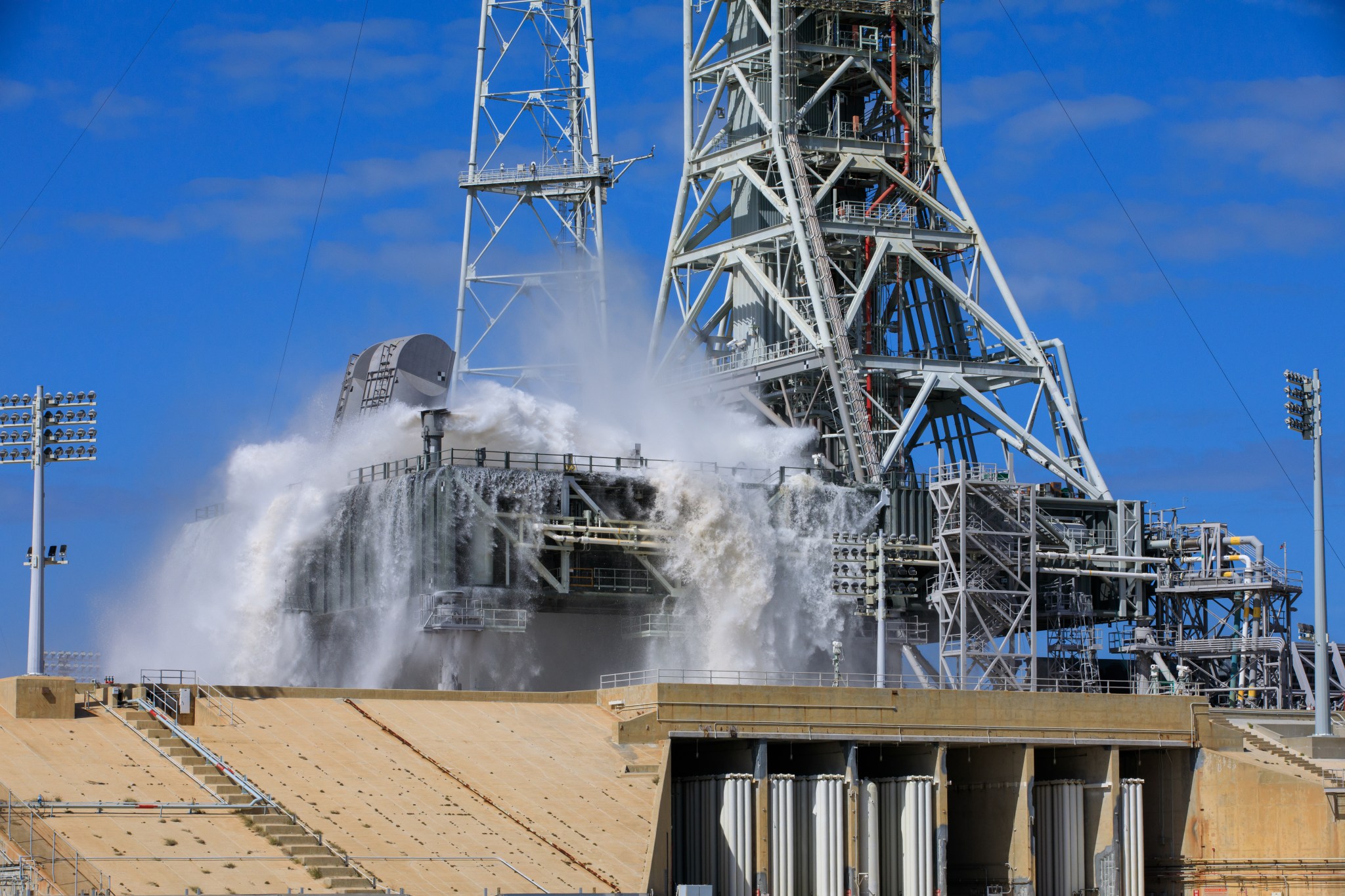 A large amount of water cascades over the edges of the gray mobile launcher at NASA's Kennedy Space Center. Droplets of water also spray up through the air, creating a mist.