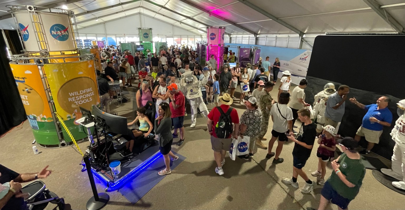 Many people wander through a large, enclosed tent. They stop to view or experience aeronautics-and space related-interactive displays.