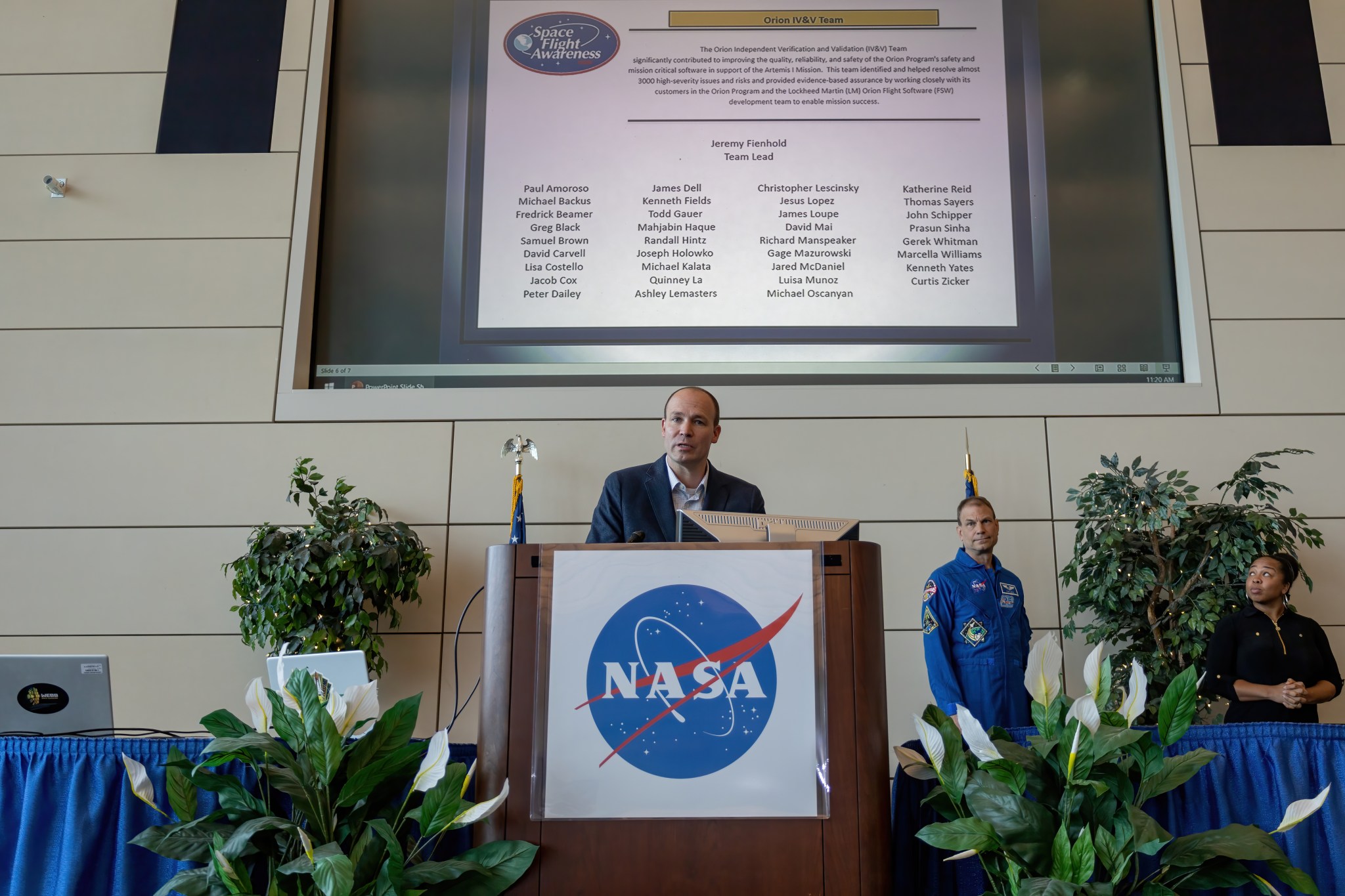 Man speaking behind a podium with a NASA logo on the front of the podium. A man in a blue jumpsuit is standing beside him. A large screen is behind them with award winners names.