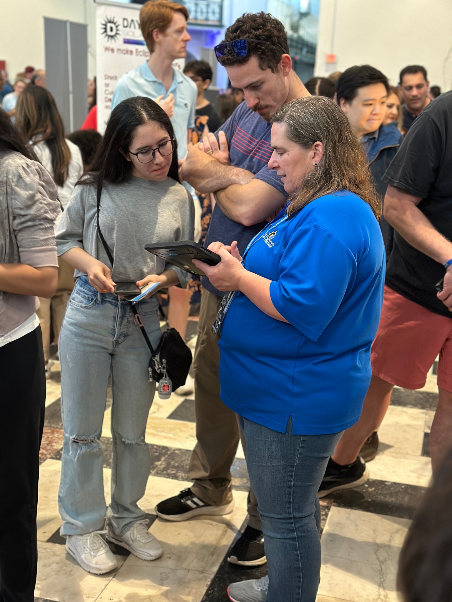 Lynn Bassford, a woman with wavy gray-brown hair, holds a tablet and speaks with members of the public at an event. She wears a bright blue shirt and jeans, and speaks to people in casual clothes who look intently at the tablet.