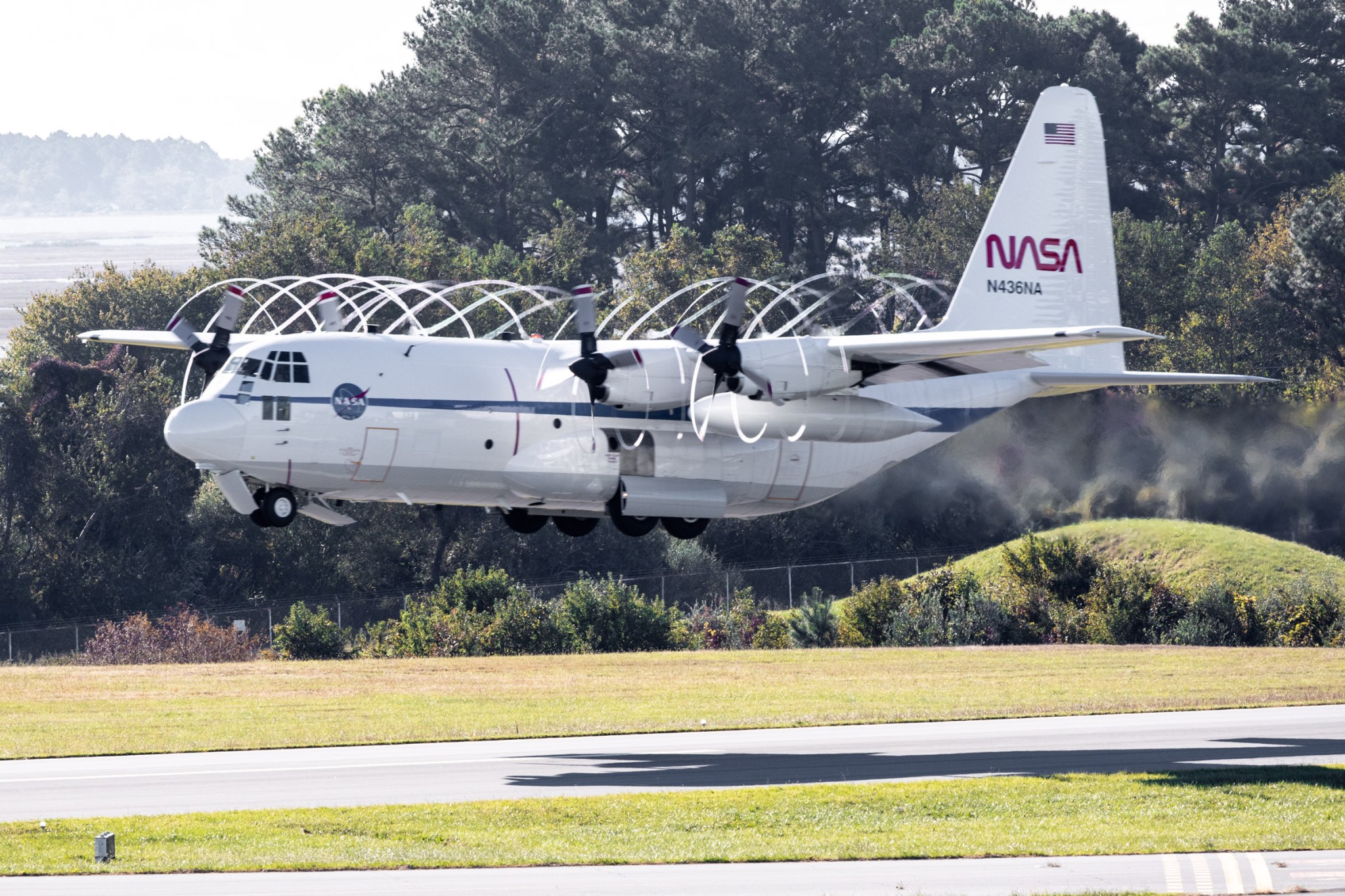 The C-130 aircraft, a white plane with a blue strip down the side, taking off from a runway with a thick tree line in the background.