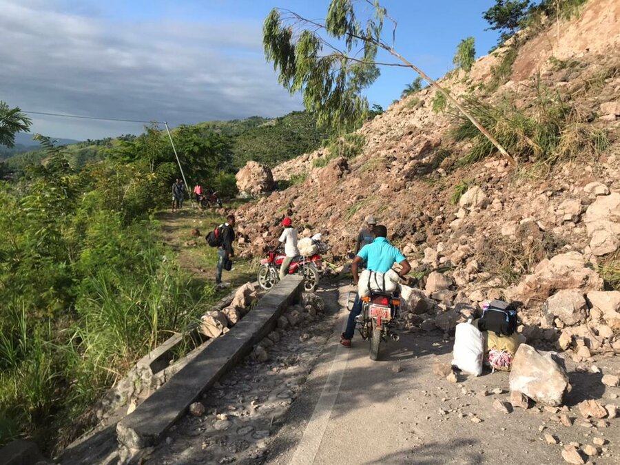 A man on a motorcycle is blocked by a landslide that has fallen across the road, covering it in large boulders, rocks and debris. A few other men working their way around the blockage. The sky is blue and slightly cloudy, and they are in a forested area. 