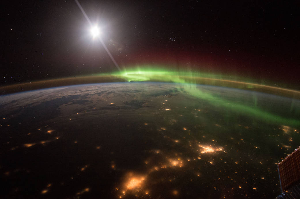 Green auroras dancing along Earth's atmosphere at night. View of city lights on Earth and the Moon in the background.