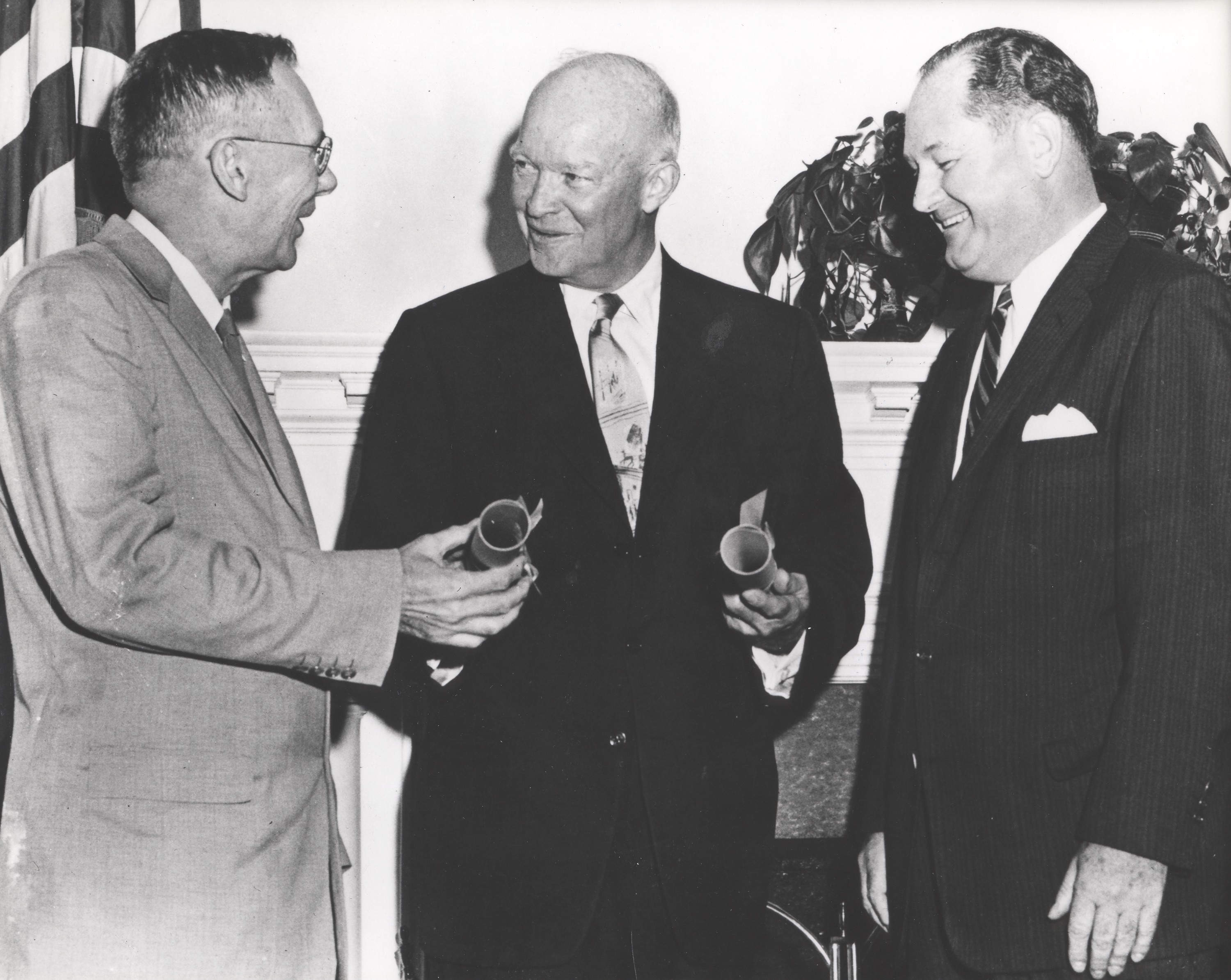 From left to right, Dr. Hugh L. Dryden, President Dwight D. Eisenhower, and Dr. T. Keith Glennan smile as they have a discussion. Dryden and Eisenhower hold cylindrical objects in their hands; Glennan looks down at their hands. They are all wearing suits.