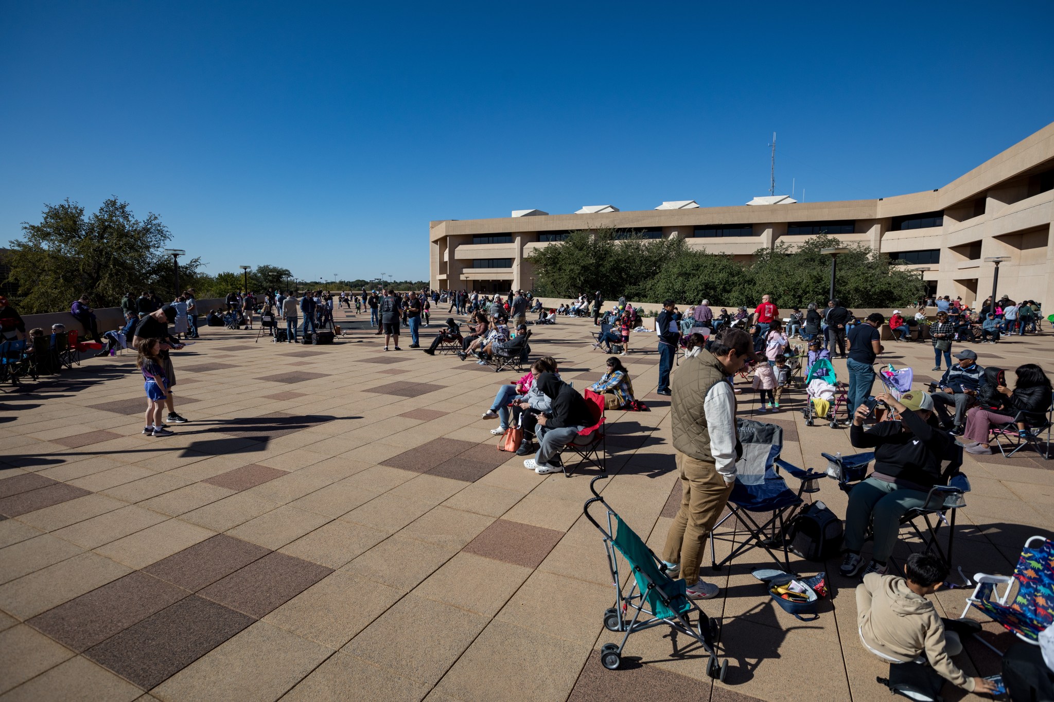 A crowd of people, including families with young children, gathers on a broad plaza paved with concrete tiles. The people are in small groups, some with lawn chairs, some sitting on the tiles. The sky is a cloudless blue.