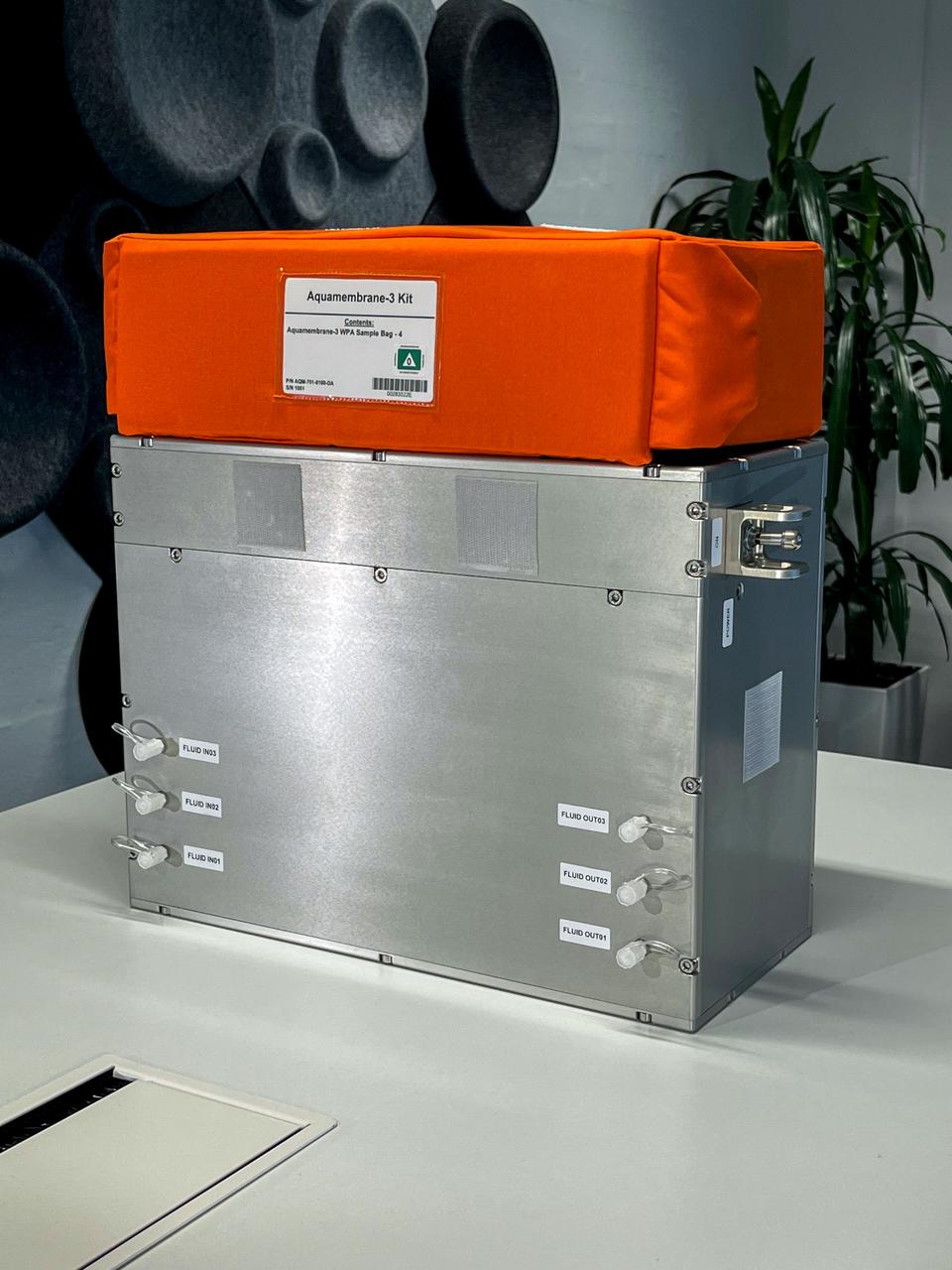 Hardware for Aquamembrane-3, a silver suitcase-sized block with six plastic fluid inputs, with a bright orange sample bag bearing a label sitting on its top.