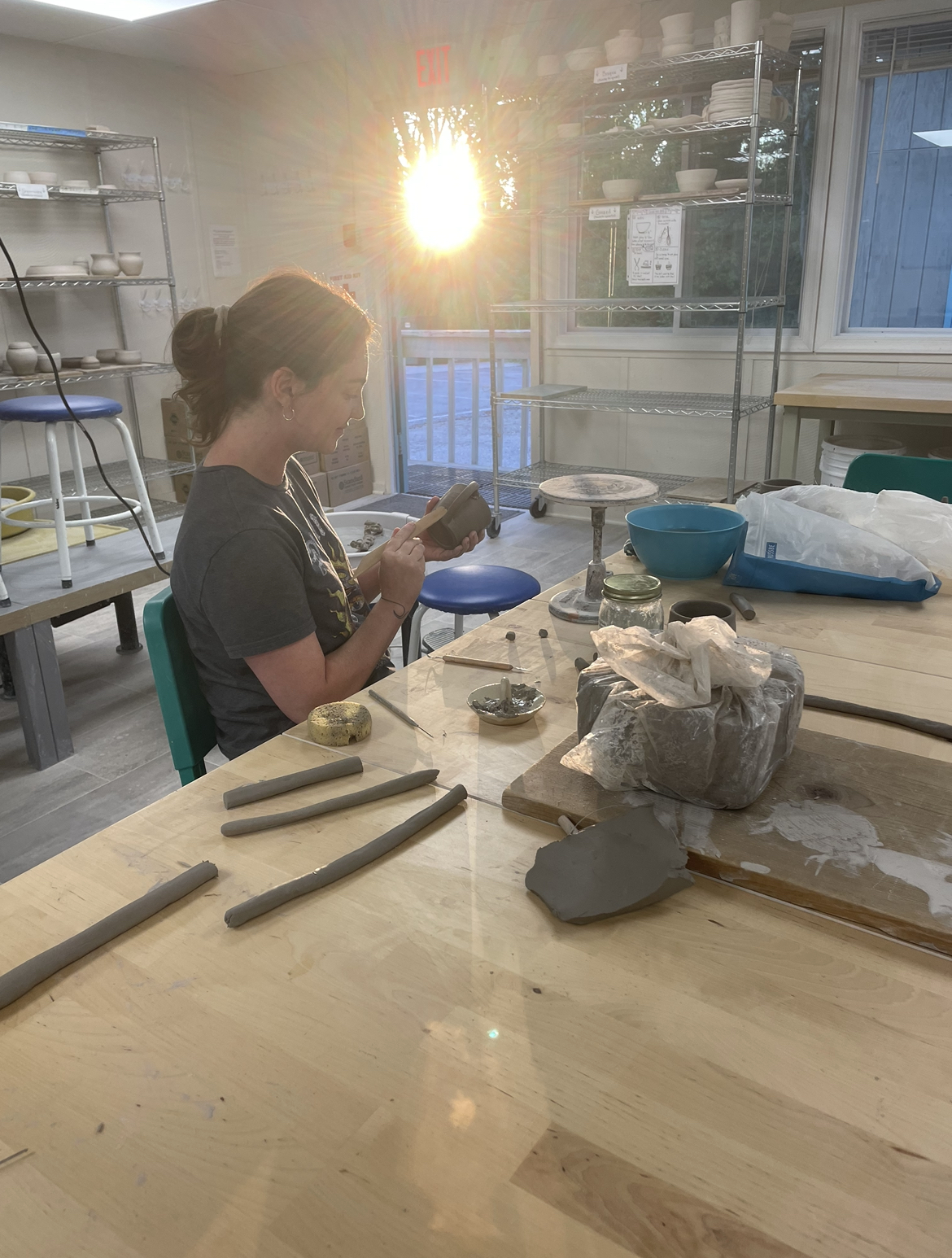 Shaigh Sisk, a woman with curly brown hair pulled back in a ponytail, sits at a pottery studio workbench sculpting a ceramic mug, while the sun shines brightly in through the studio door and casts rays across the image. Shaigh wears a dark gray tee and small hoop earrings. The mug in her hands is gray clay, and blocks and rolls of the same clay sit on the bench near her, along with sculpting tools and sponges. Racks with drying pottery in various shades of beige are visible behind her.