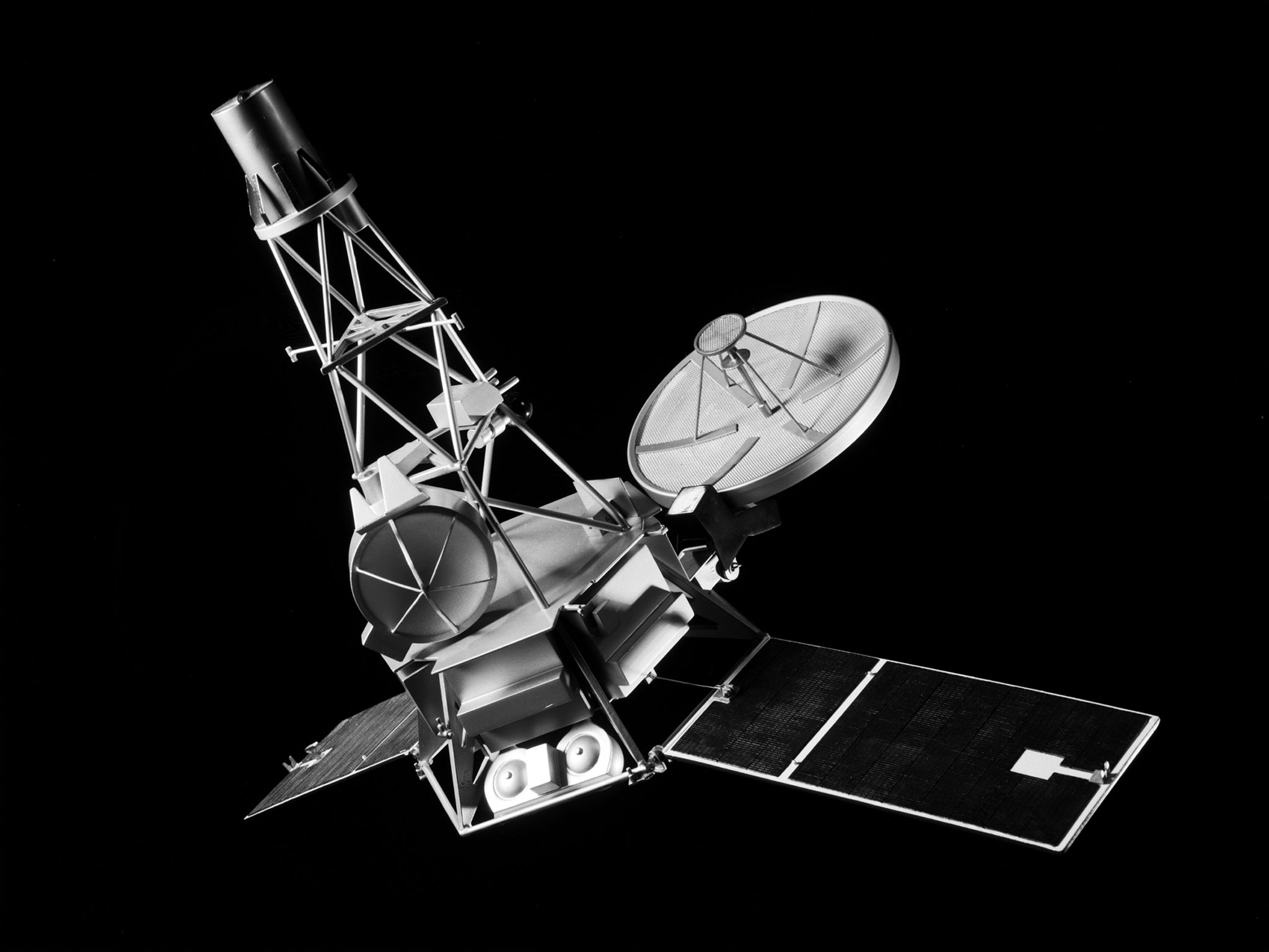 A black and white photo of a model of the Mariner-C spacecraft against a black backdrop. The spacecraft is tilted to the left. It has a conical shape, with two solar panels and a satellite dish at the bottom.