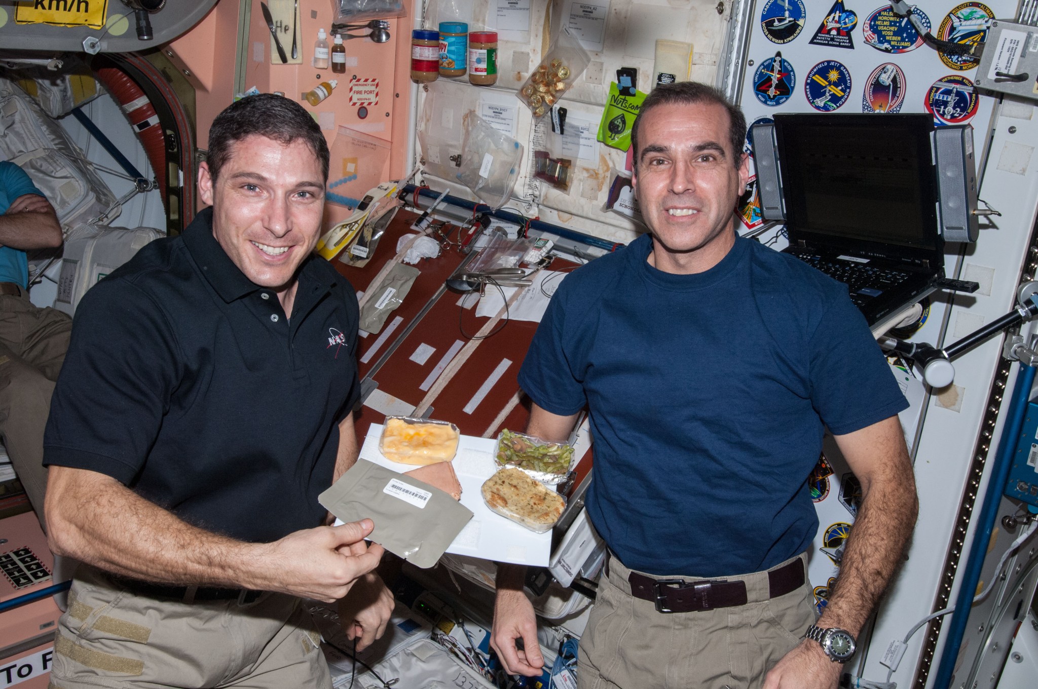 NASA astronauts Michael Hopkins (left) and Rick Mastracchio smile for the camera while aboard the International Space Station. Hopkins holds a thin white sheet with Velcro on it, which secures the Thanksgiving foods shown, including green beans, smoked turkey, and stuffing.