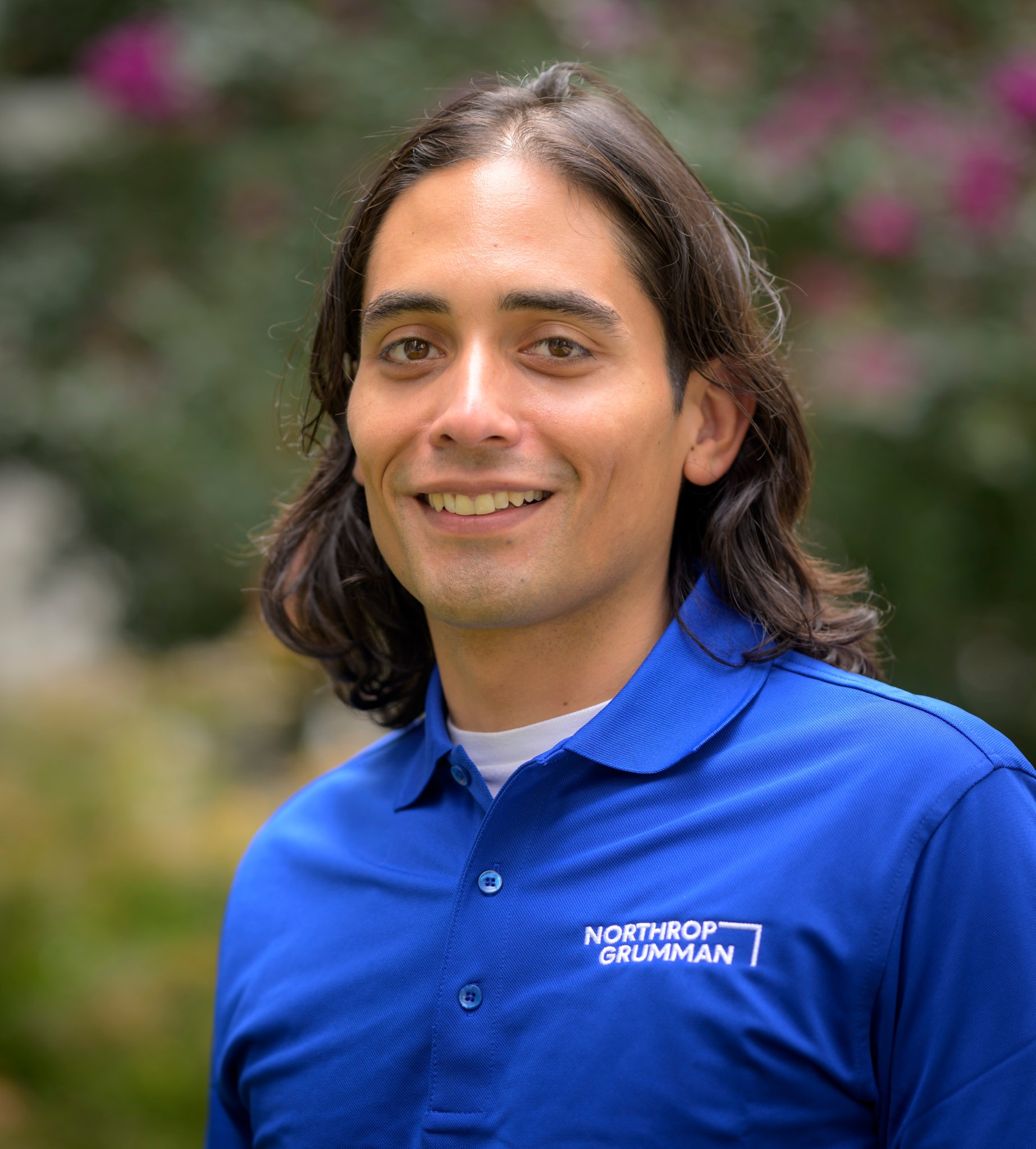 A portrait of Northrop Grumman engineer Oliver Ortiz, who has shoulder-length brown hair, is wearing a blue shirt with the Northrop Grumman logo on the left breast, as he flashes a smile and stands in front of some green and magenta-speckled foliage.