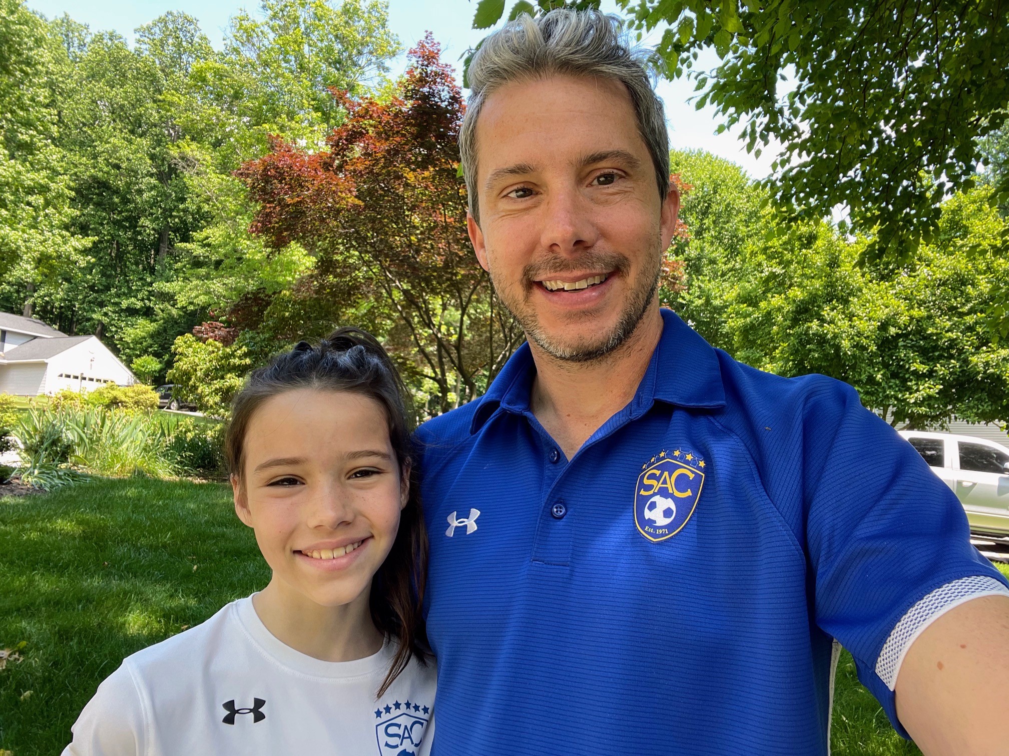 Joshua Abel, a man with short gray hair and a short dark gray beard, smiles and poses with his daughter for a selfie. Joshua wears a bright blue soccer polo and his daughter, a young girl with long dark hair, wears a white soccer jersey. They pose in the shade of a large tree, with yards, driveways and more trees visible behind them.