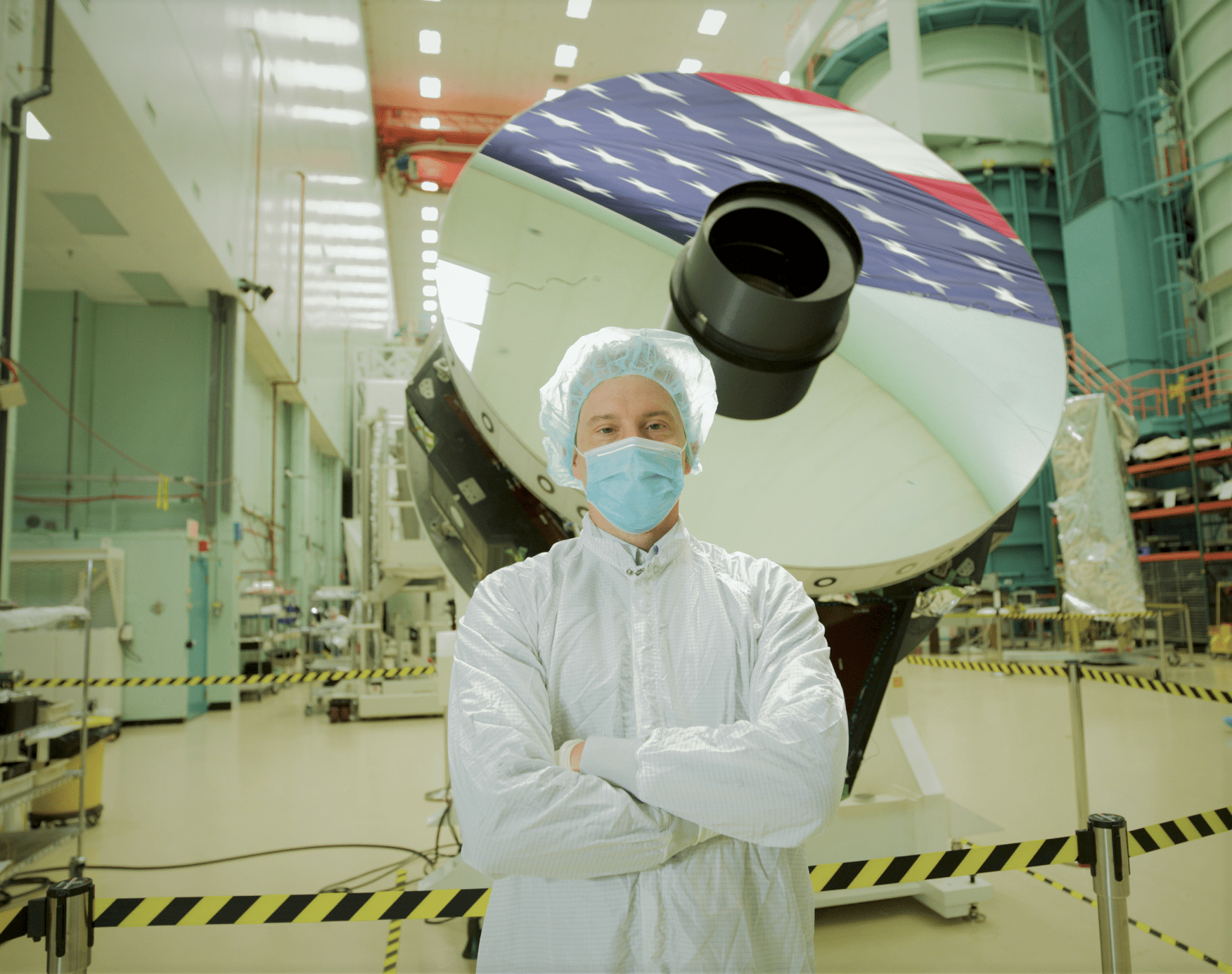Joshua Abel, a man wearing white coveralls, a light blue hair net, and a light blue face mask, stands and poses with arms crossed in front of the Nancy Grace Roman Space Telescope's primary mirror. The mirror is shaped like a large silver disk, reflecting part of an American flag in its upper surface. Both Joshua and the mirror are inside a clean room, with pipes, shelves, stairs, and storage lining the walls, most in shades of light turquoise. Black and yellow caution tape forms a barrier around the telescope mirror.