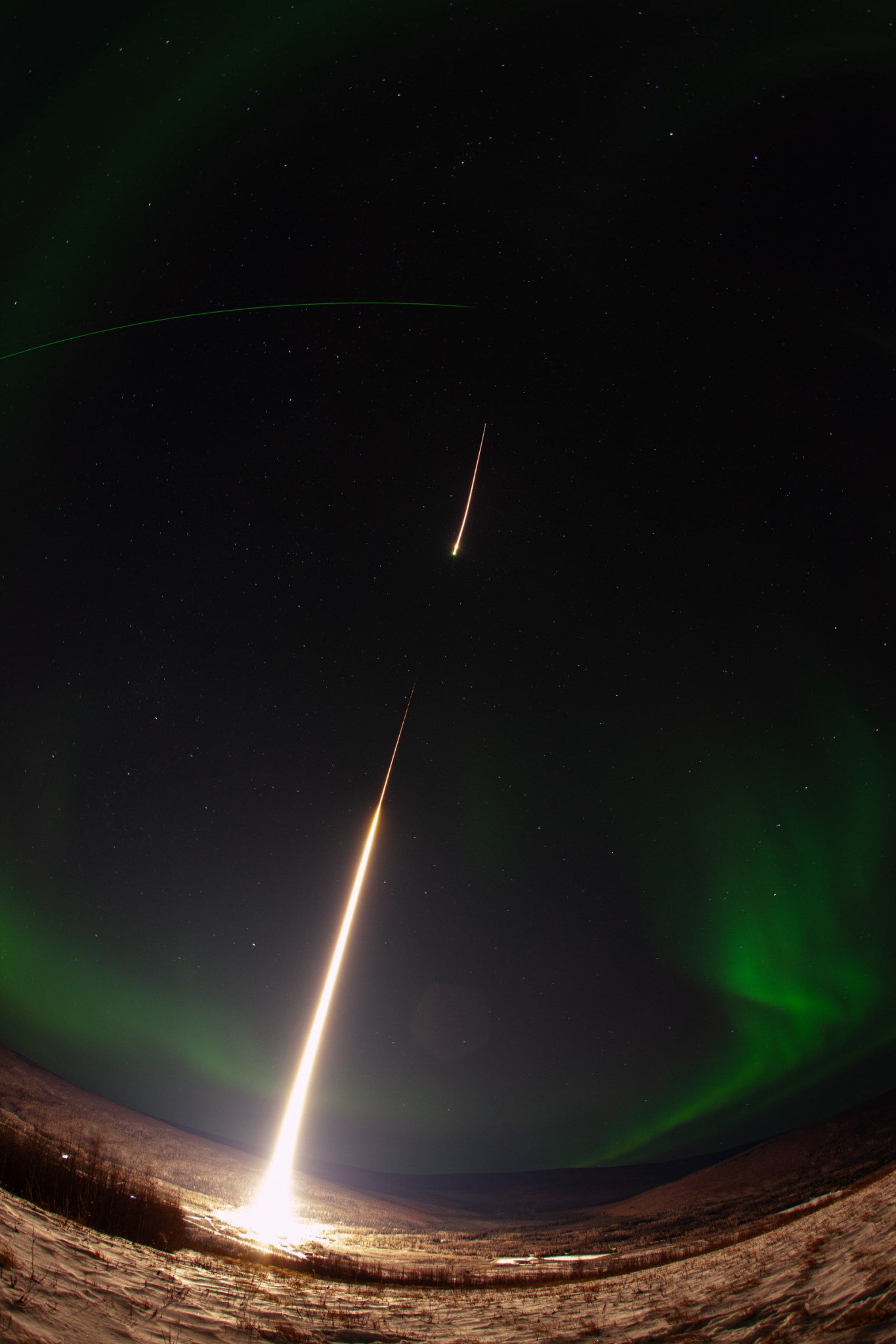 A long-exposure photograph of a sounding rocket launching in a night sky highlighted by aurora. The sounding rocket is a bright white streak, leaving from a snow-covered ground and moving up into the sky. A small break in the streak represents the first stage of the rocket burning out and the second stage igniting. A soft, green aurora frames the edges of the image, with several white stars speckled through the black sky. A bright green line toward the top of the frame represents a LIDAR beam. A fisheye lens was used for the photograph, creating a curve for the ground and LIDAR beam.