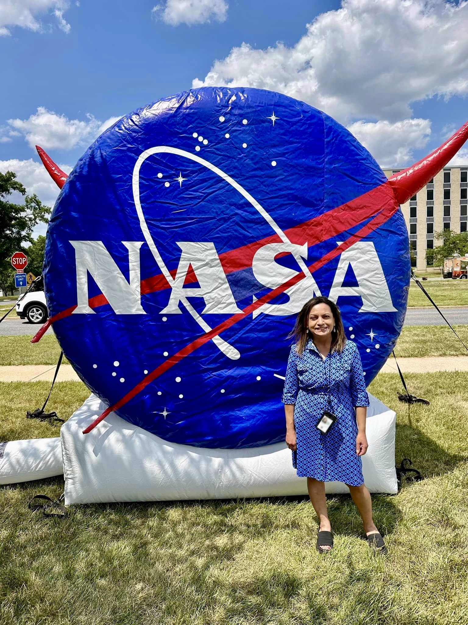Rita Owens, a woman with shoulder-length, light brown hair, smiles and stands in front of a large inflatable shaped like the NASA logo. It is a large, bright blue circle dotted with stars and crossed by a red, V-shaped swoosh. "NASA" is written in large white text on the circle. Rita wears a patterned blue dress and black sandals. It is a bright, sunny day with blue sky, puffy clouds, and green grass. A building and trucks are visible behind the NASA inflatable.