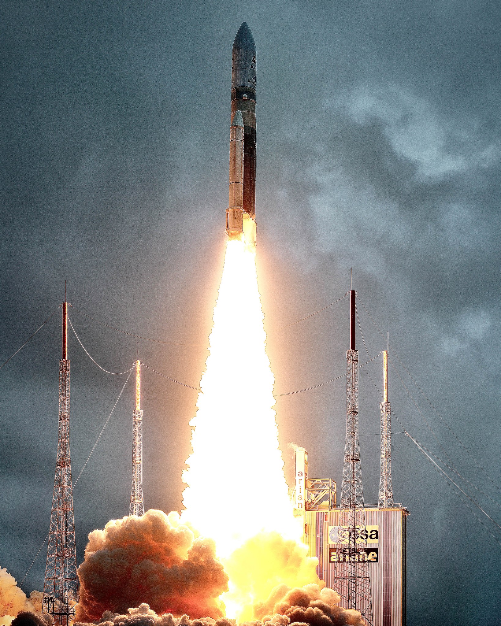 Webb’s Ariane 5 rocket launches from Europe’s Spaceport in Kourou, French Guiana. A glowing, white-yellow stream of gas and fire is streaming from the rocket towards the ground below. The ground is covered with puffy clouds of pink-orange gas. In the background, lit up by the launch, is a building that is emblazoned with dark blue European Space Agency (ESA) and Ariane logos. The sky is a dark blue-gray and largely cloudy.