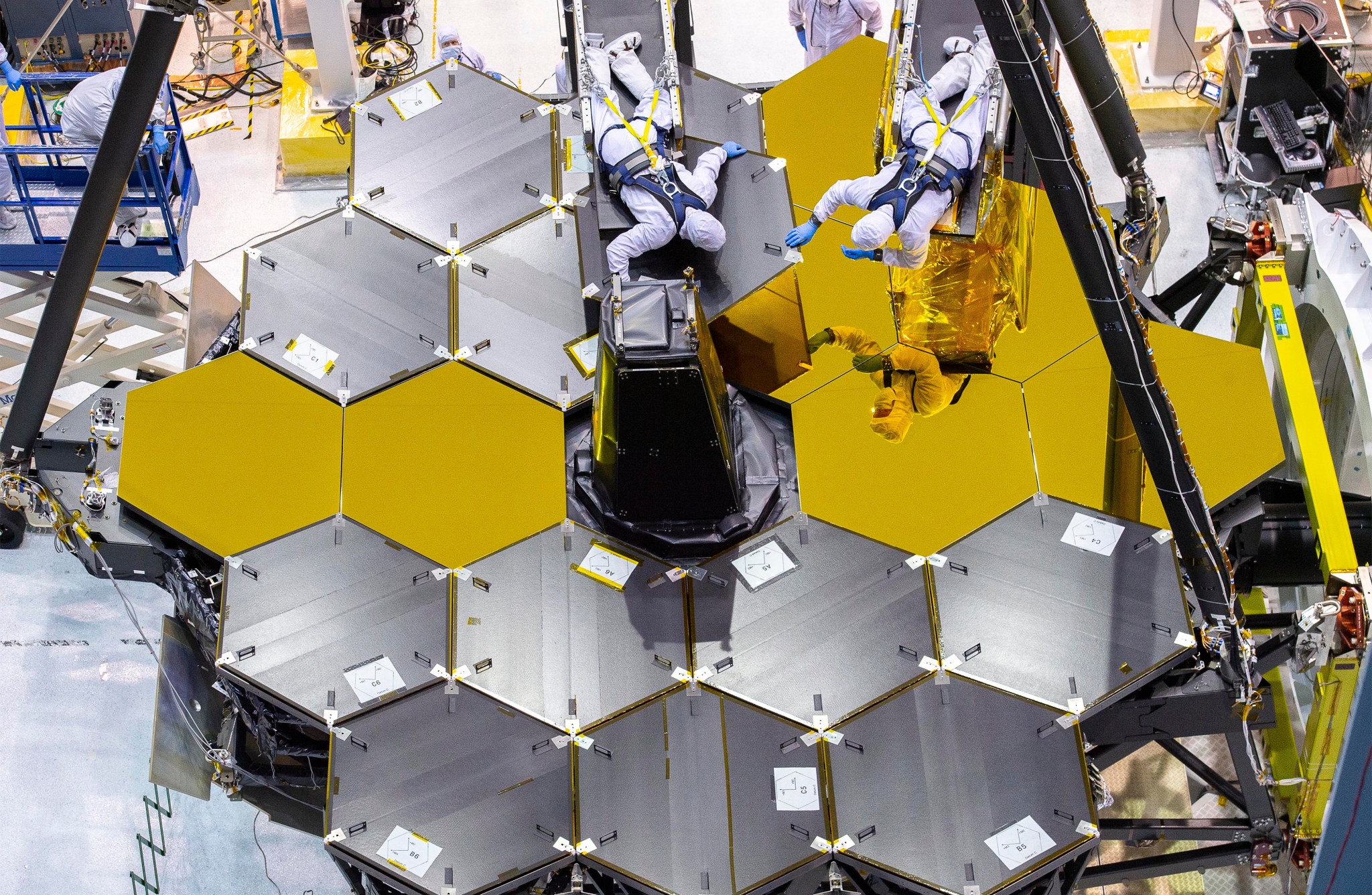 The covers being removed from the hexagonal gold mirrors of the James Webb Space Telescope. Two people in cleanroom “bunny” suits lay on diving boards over the mirror, which lays face pointed up, very carefully removing the covers one at a time. In this photo 6 covers have been removed, revealing the golden mirrors beneath. These are the 4 center line mirrors and two in the 4 and 5 o’clock positions. The two workers each hold part of another cover as they remove it. The worker on the right is reflected in the mirrors. Each of the black covers has a white sign on it noting which mirror segment it is. Each segment has a unique designation including A, B, or C and 1, 2, 3, 4, 5, or 6 depending on its prescription and location.