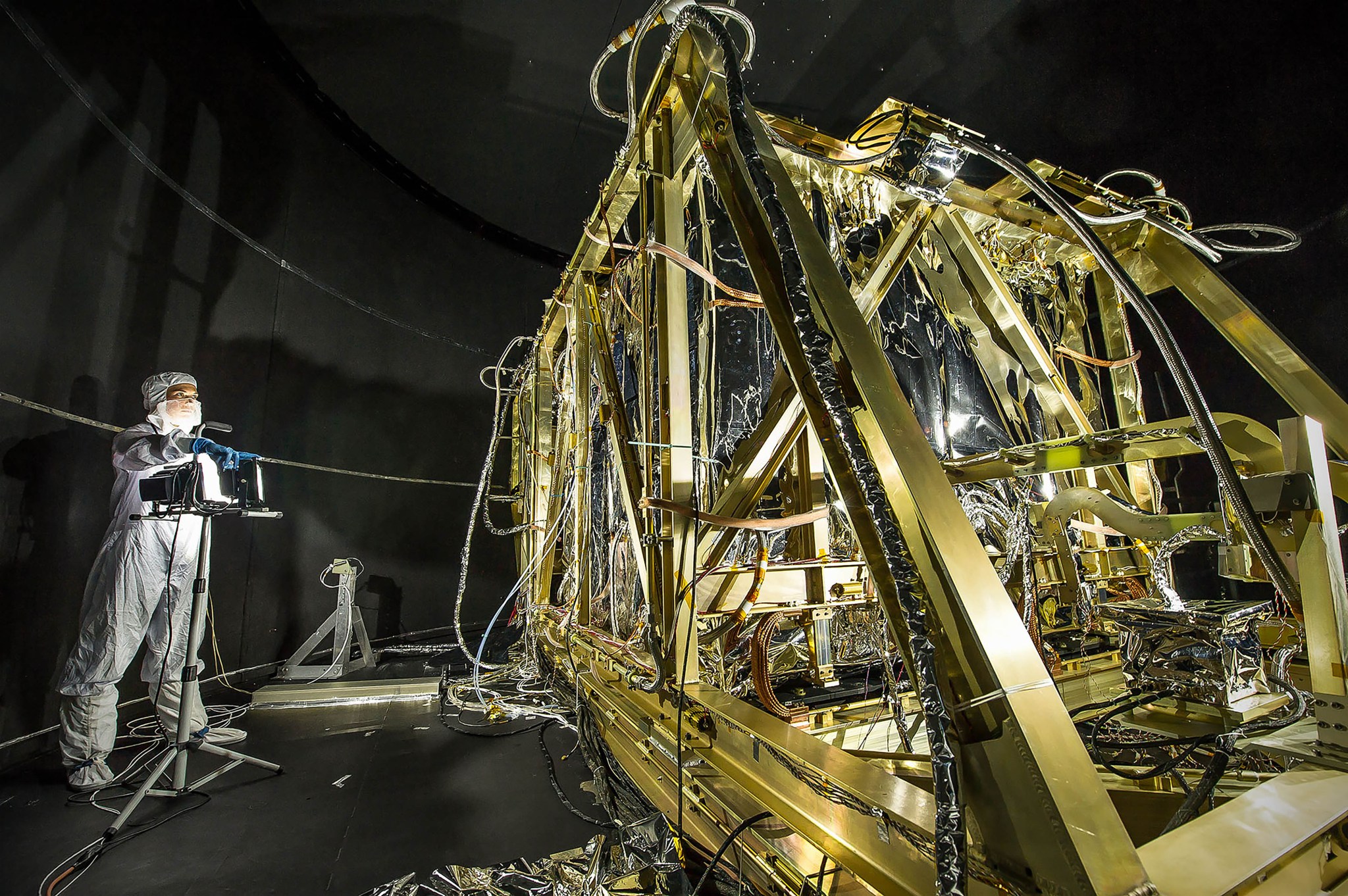 The silvery James Webb Space Telescope instrument module is enclosed in a metallic gold-colored structure. NASA photographer Desiree Stover stands to the left, holding some equipment and facing the hardware. She is wearing a white cleanroom suit. She and the hardware are inside a round, black chamber, which is inside Goddard’s large thermal vacuum chamber where the instrument is tested at its extremely cold operating temperatures.
