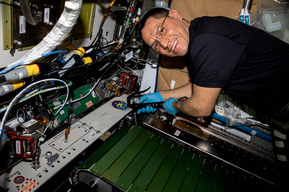 NASA astronaut Frank Rubio’s body is facing the treadmill while he turns his head to smile at the camera. He is wearing blue sterile gloves while holding a tool. Above Rubio’s head are several wires and cords.