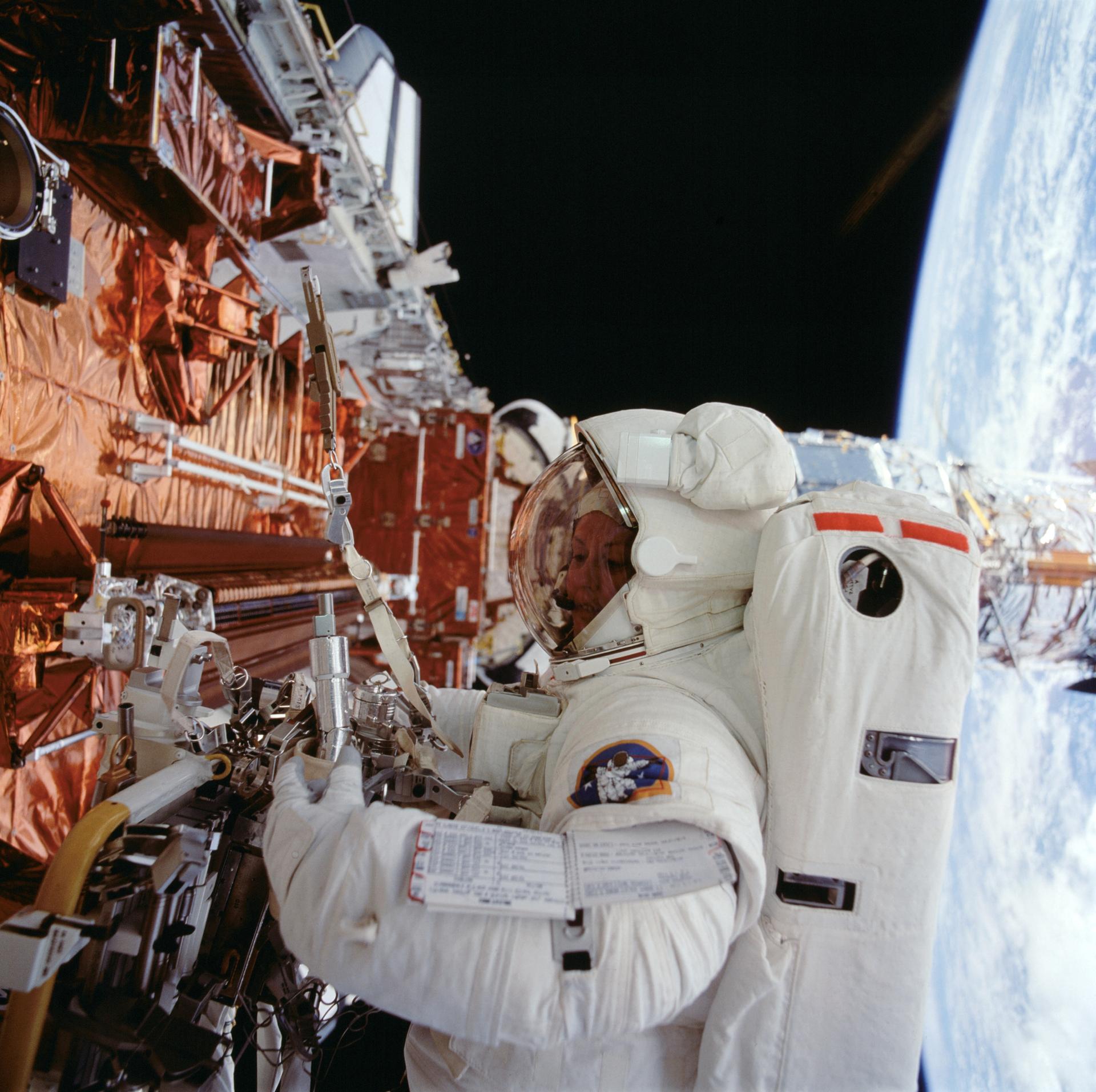 An astronaut at one of the gold covered cargo bay carriers with the earth and Hubble off to the right.