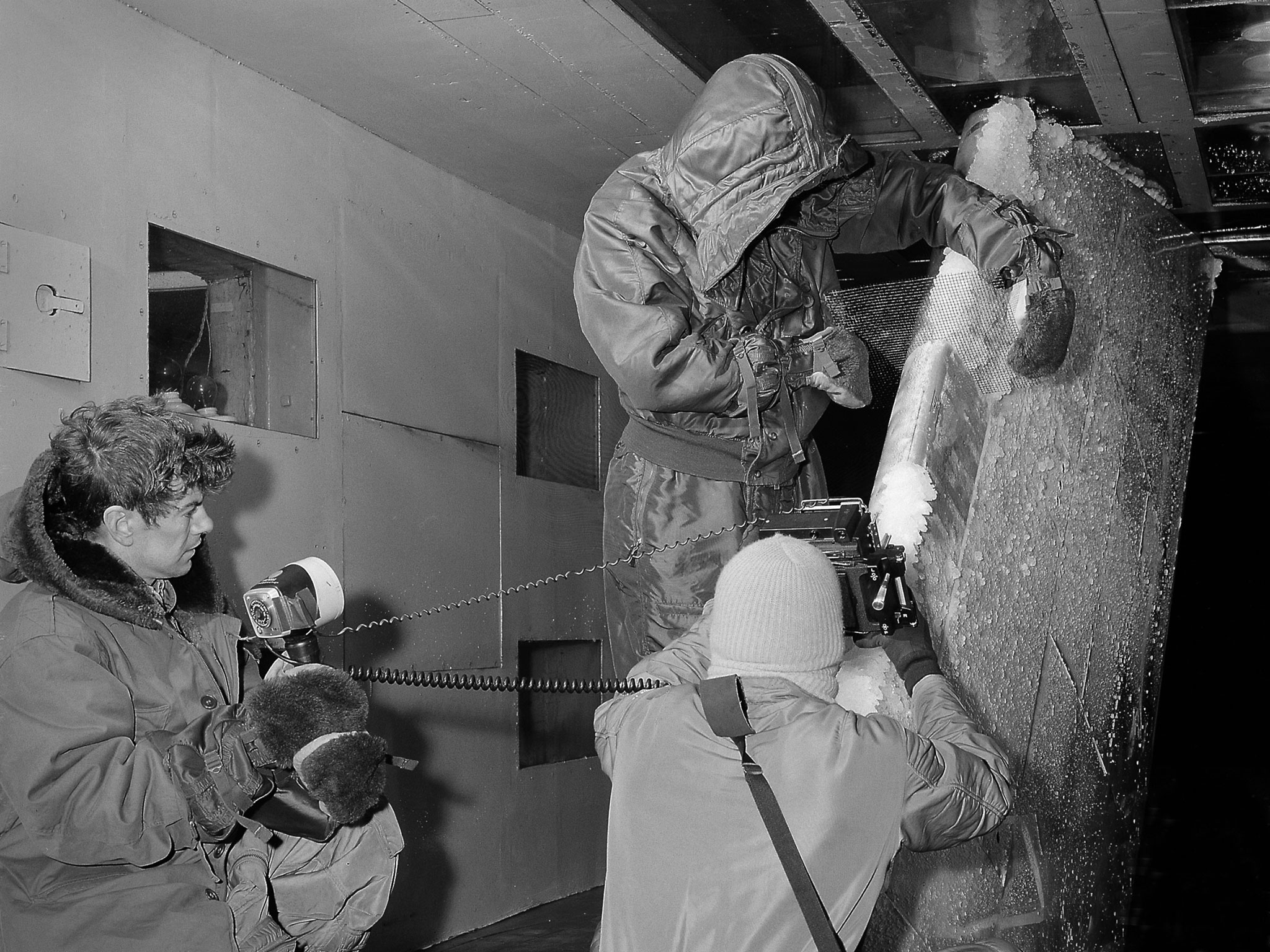 A black-and-white photo of three men dressed in winter gear working on a piece of test hardware inside NASA’s Icing Research Tunnel. All of the men are wearing coats and mittens and the one at the center also wears a hat. Chunks of ice are visible on the test hardware.