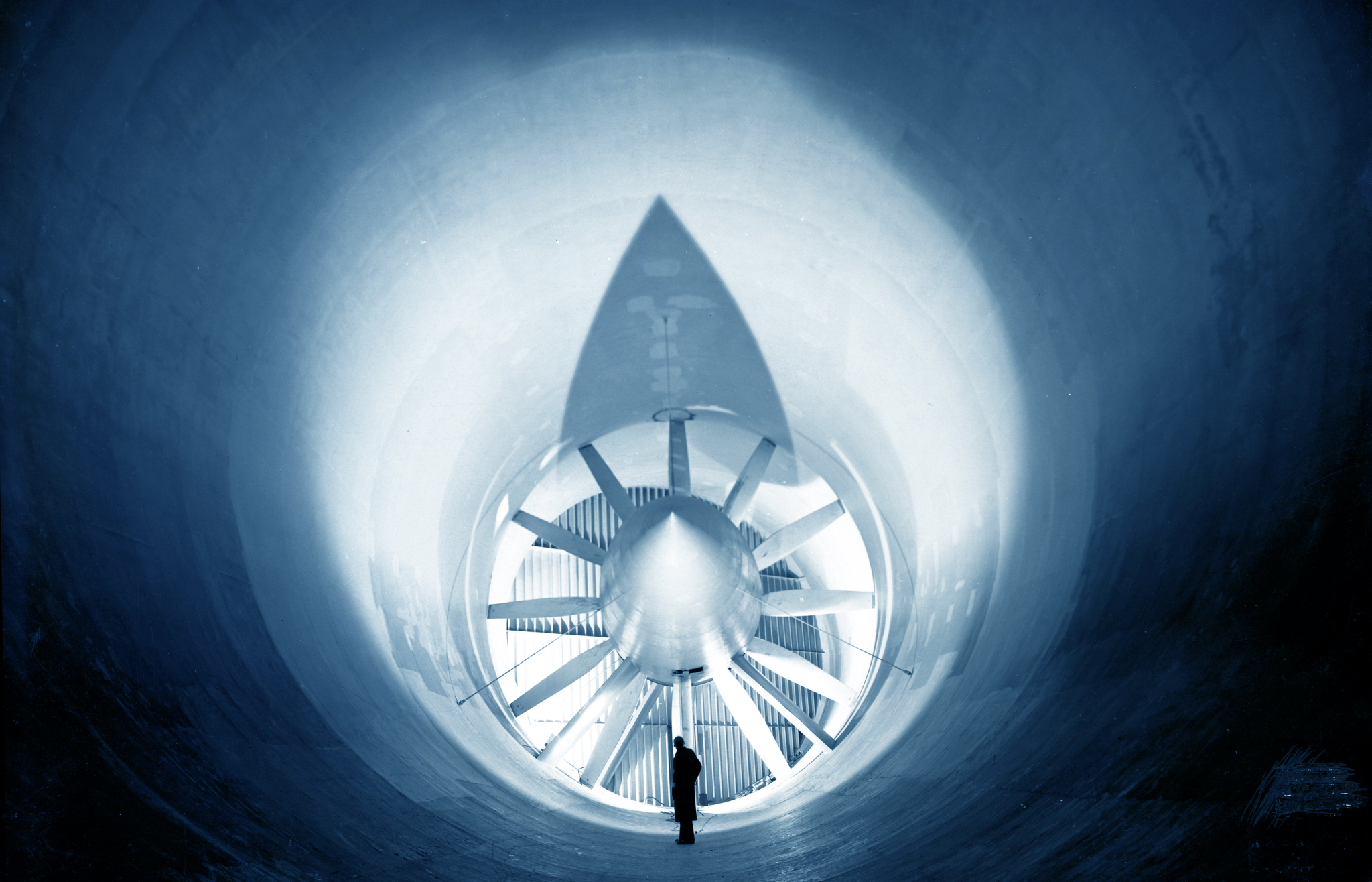 A man stands beneath the blades of a very large fan inside the Altitude Wind Tunnel. The foreground of the photo is dark, and only a silhouette of the person is visible. The area closer to the fan is illuminated.