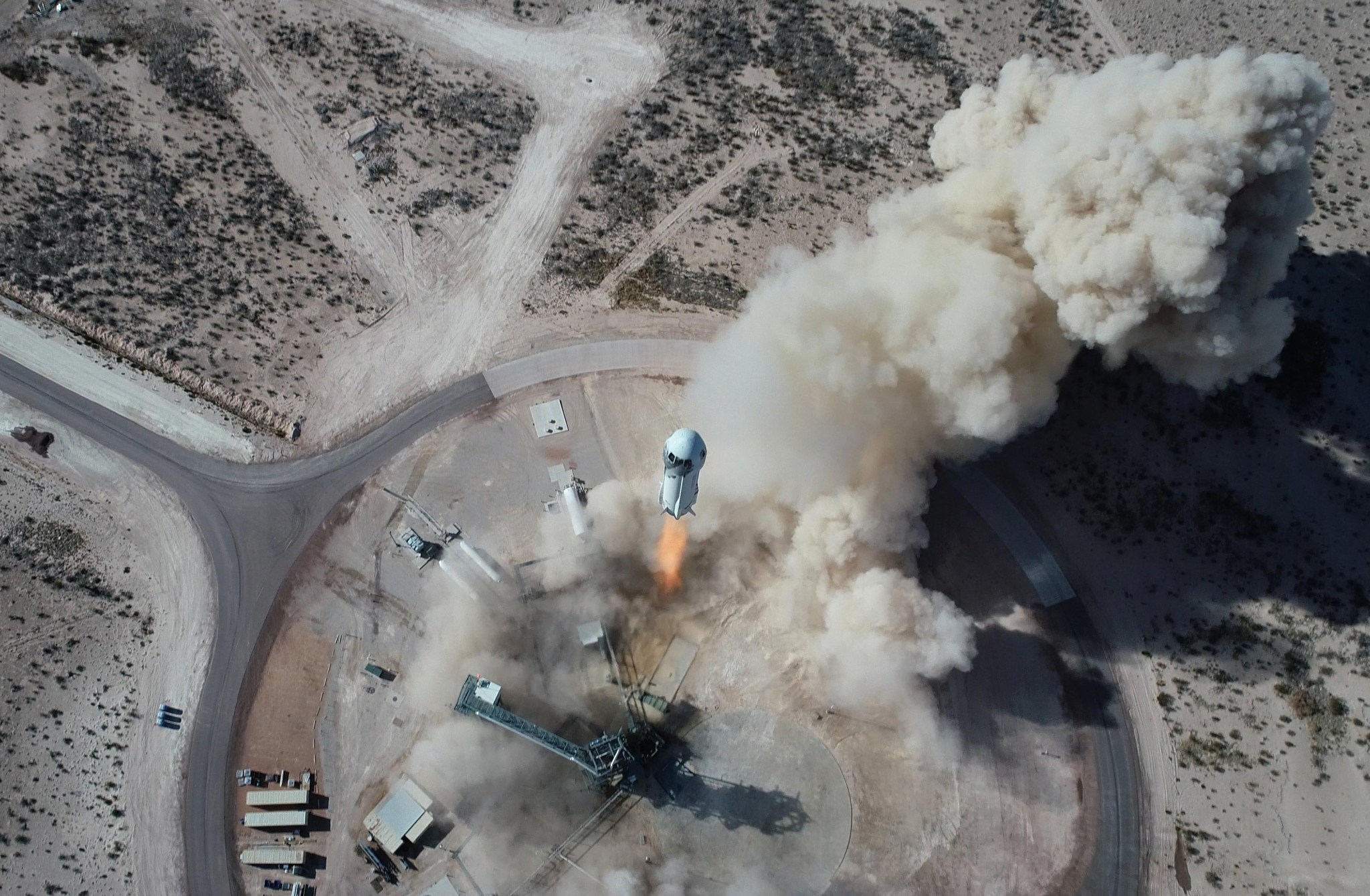 Aerial view of a desert area. Fire emerges from bottom of rocket rising from launchpad. Smoke and dust billow skyward.
