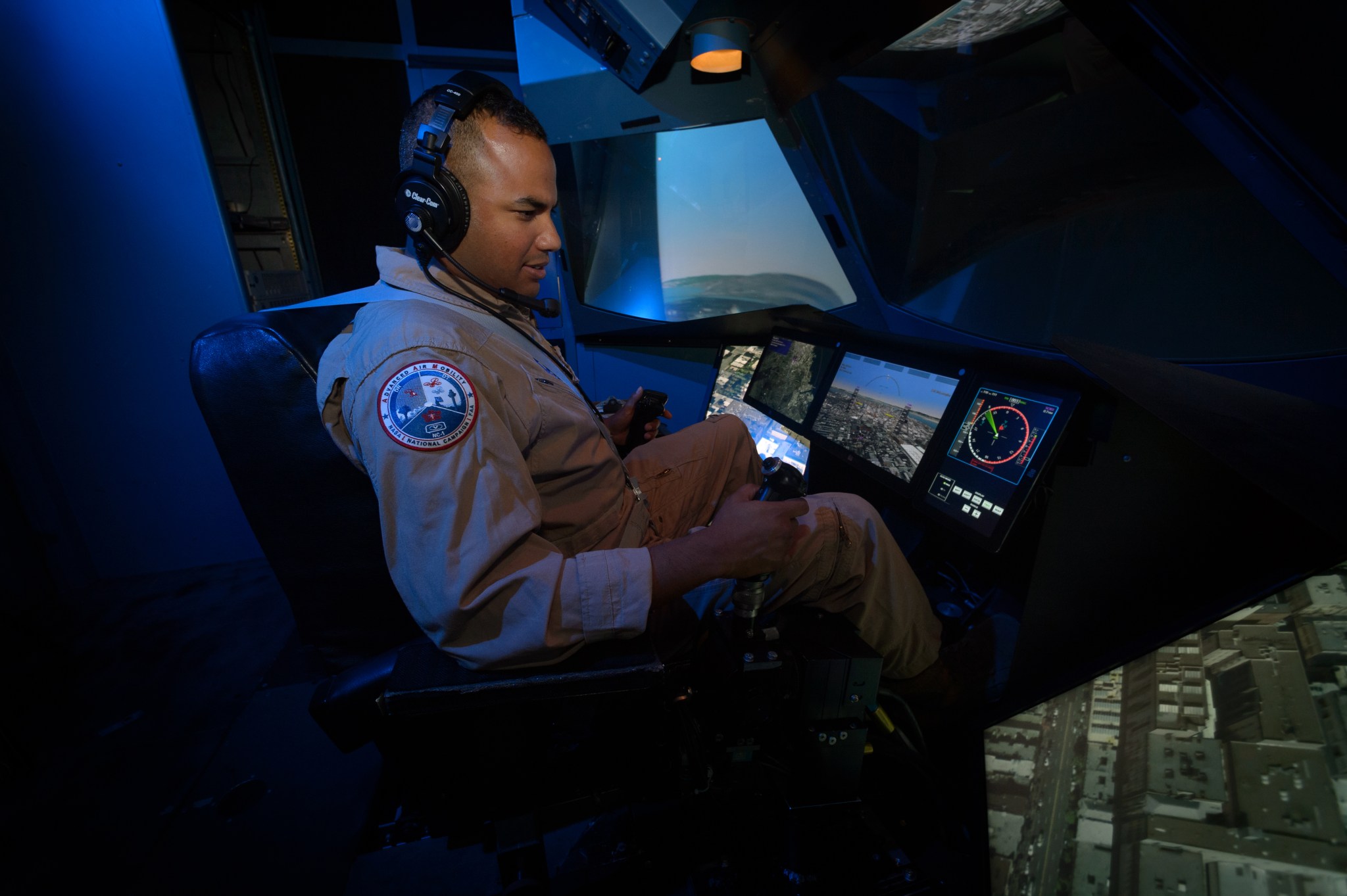 A man wearing a tan flight suit flies in a blue-lit flight simulator. Navigation guidance and other controls light up the simulator’s front dashboard.