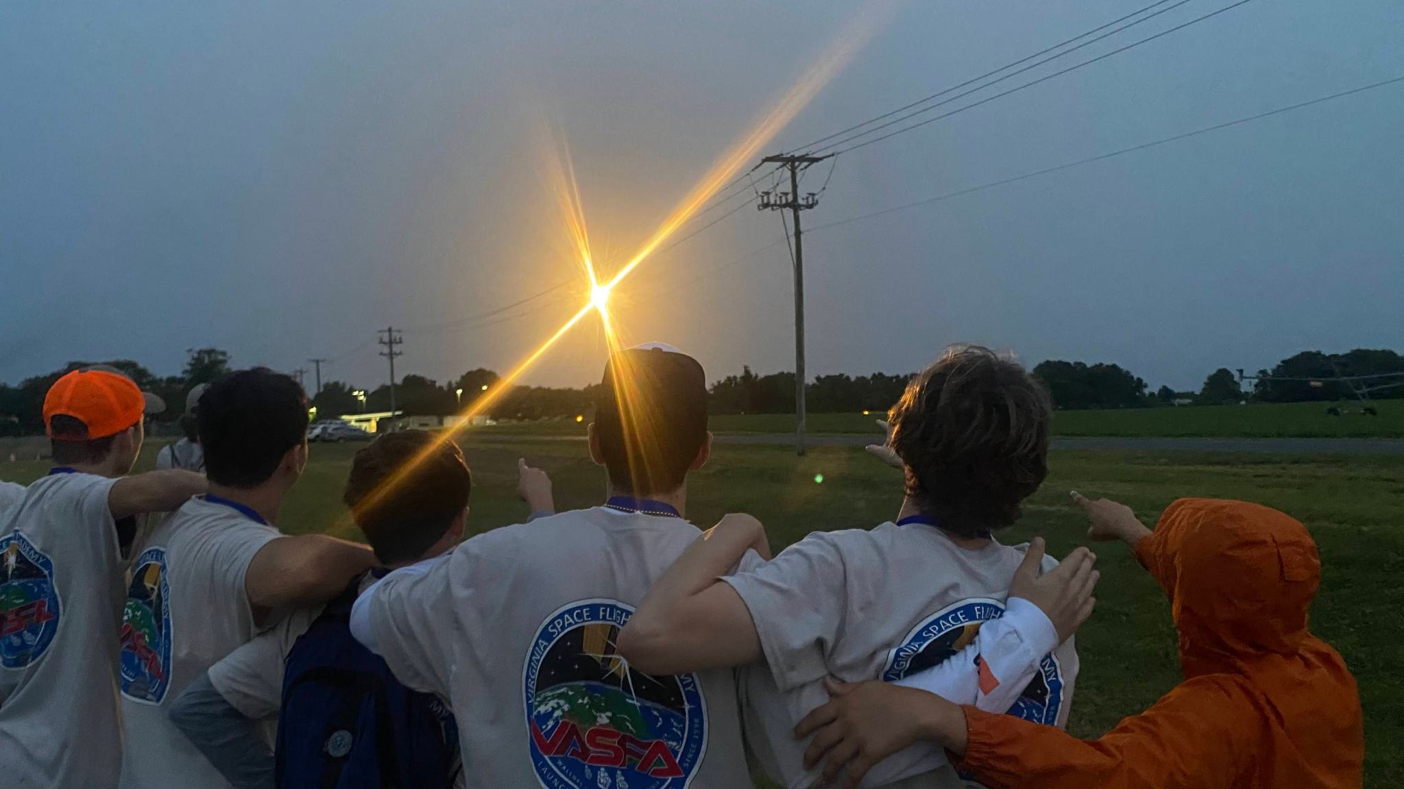 five people stand with their backs facing the camera, all five people are looking and pointing at a bright orange light, rising above the horizon.