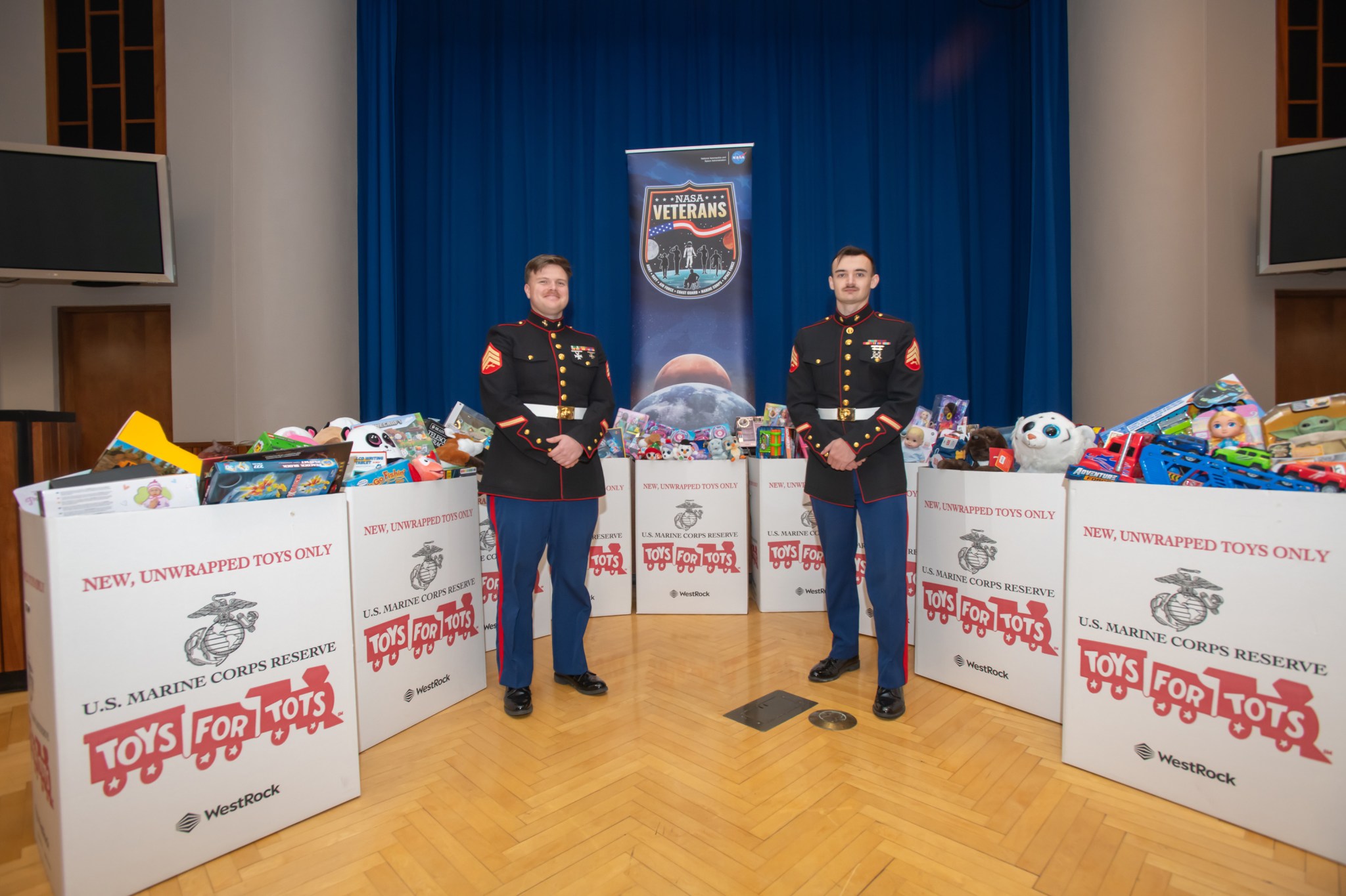 Two Marine Corps reservists in uniform stand in front of a stage with ten boxes filled with toys. The NASA Veterans emblem banner hangs high in the background.
