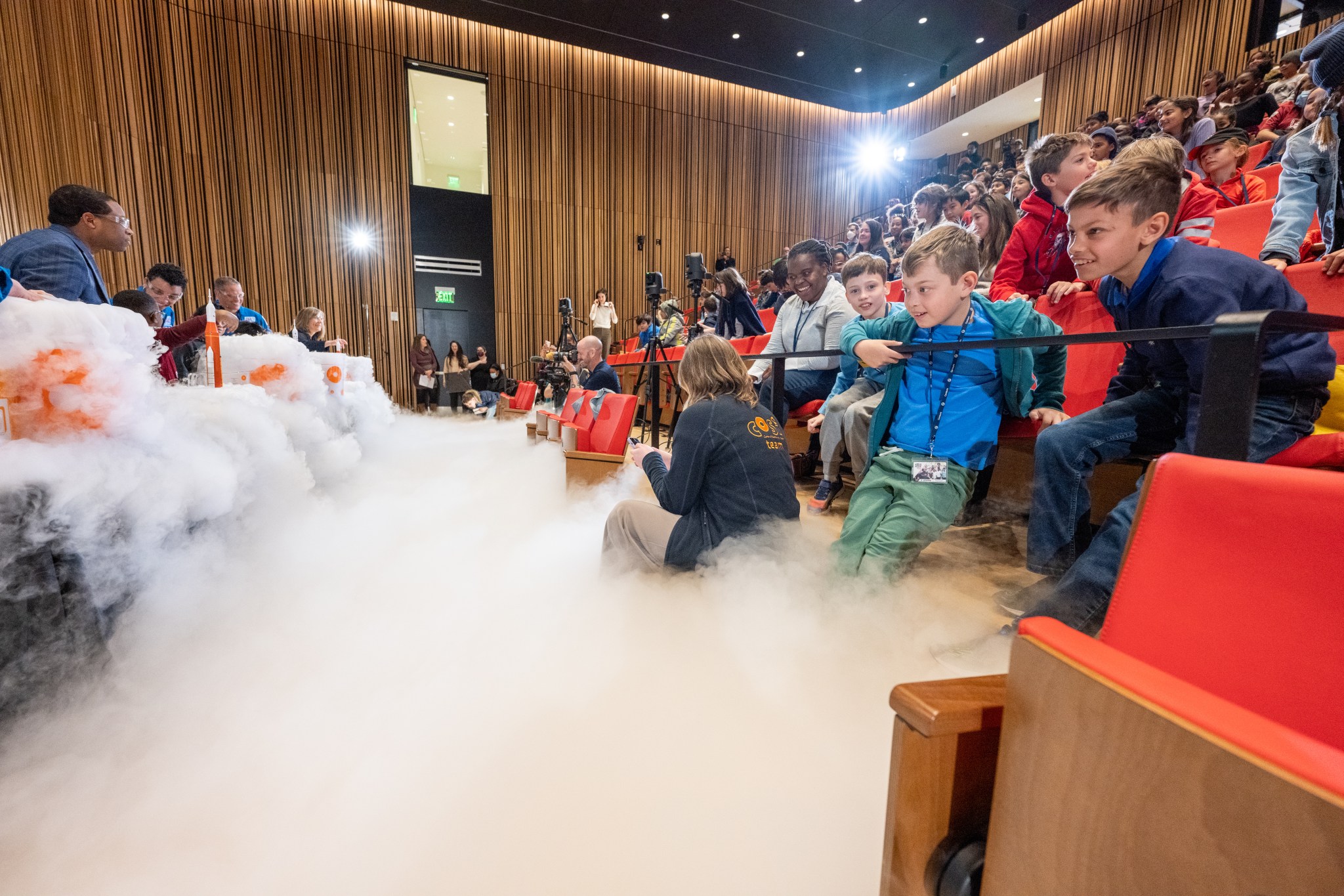 Fog rolls across the floor of an auditorium full of students and teachers. In the background at left are NASA astronauts Jessica Watkins and Bob Hines, along with students and teachers. To the right are rows of orange seats, where the audience sits, watching the experiment underway.