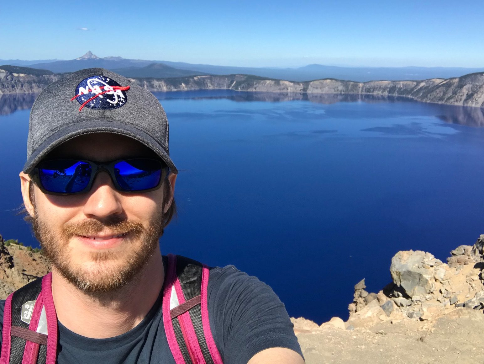 Joshua Schlieder, a man with a short brown beard, smiles and holds the camera up in a selfie outdoors near Crater Lake, Oregon. Joshua wears blue reflective sunglasses, a gray baseball cap with the NASA logo, and a navy T-shirt, and carries a backpack with pink straps. He takes up part of the left side of the image, with the still, deep blue lake visible in the background. Tan rocks and dirt are visible in the foreground, and the lake's rocky opposite shore is visible in the distance, with blue mountains on the horizon. It is a bright sunny day and the sky is cloudless and blue.