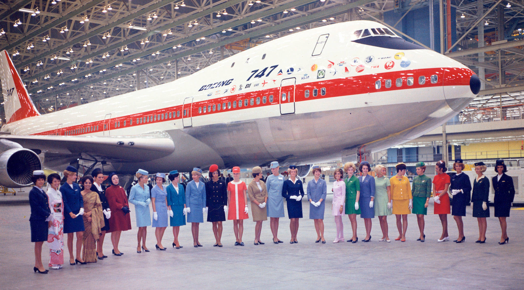 A Boeing 747 Jumbo Jet is seen in a hangar with 26 flight attendants representing 26 airliner standing in front of the airplane.