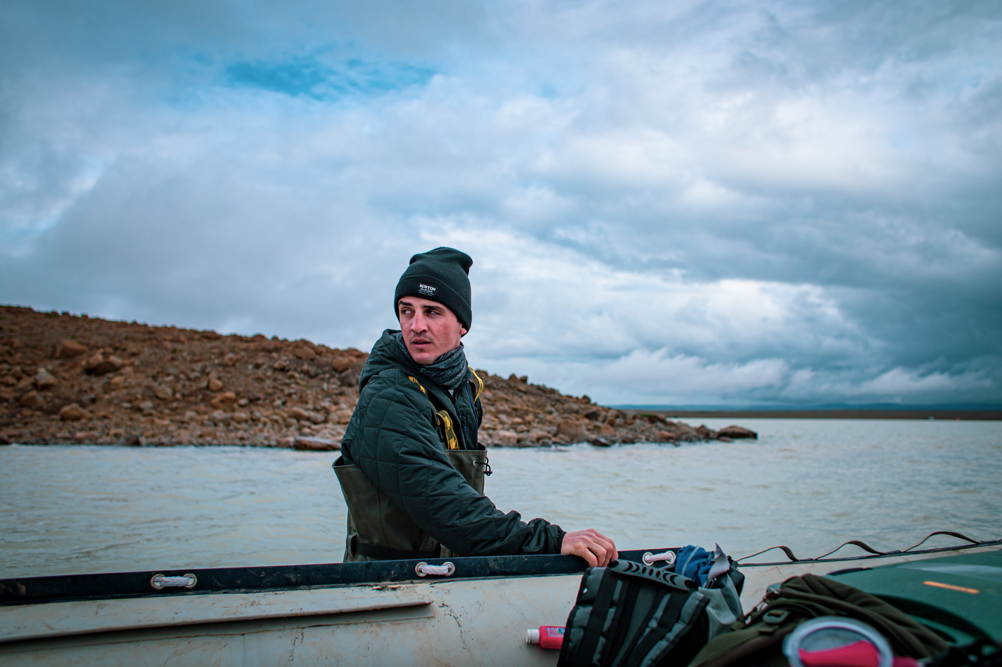 Dr. Michael Thorpe turns to look over his shoulder as he stands in water against a cloudy gray-blue sky. He wears a dark green stocking cap and a dark green quilted jacket underneath dark green waders, or waterproof overalls. His right hand rests on the side of a small boat with backpacks sitting on its top. A rocky brown coastline extends into the image behind him, and the clouds hang low over a dark blue horizon far off in the distance.