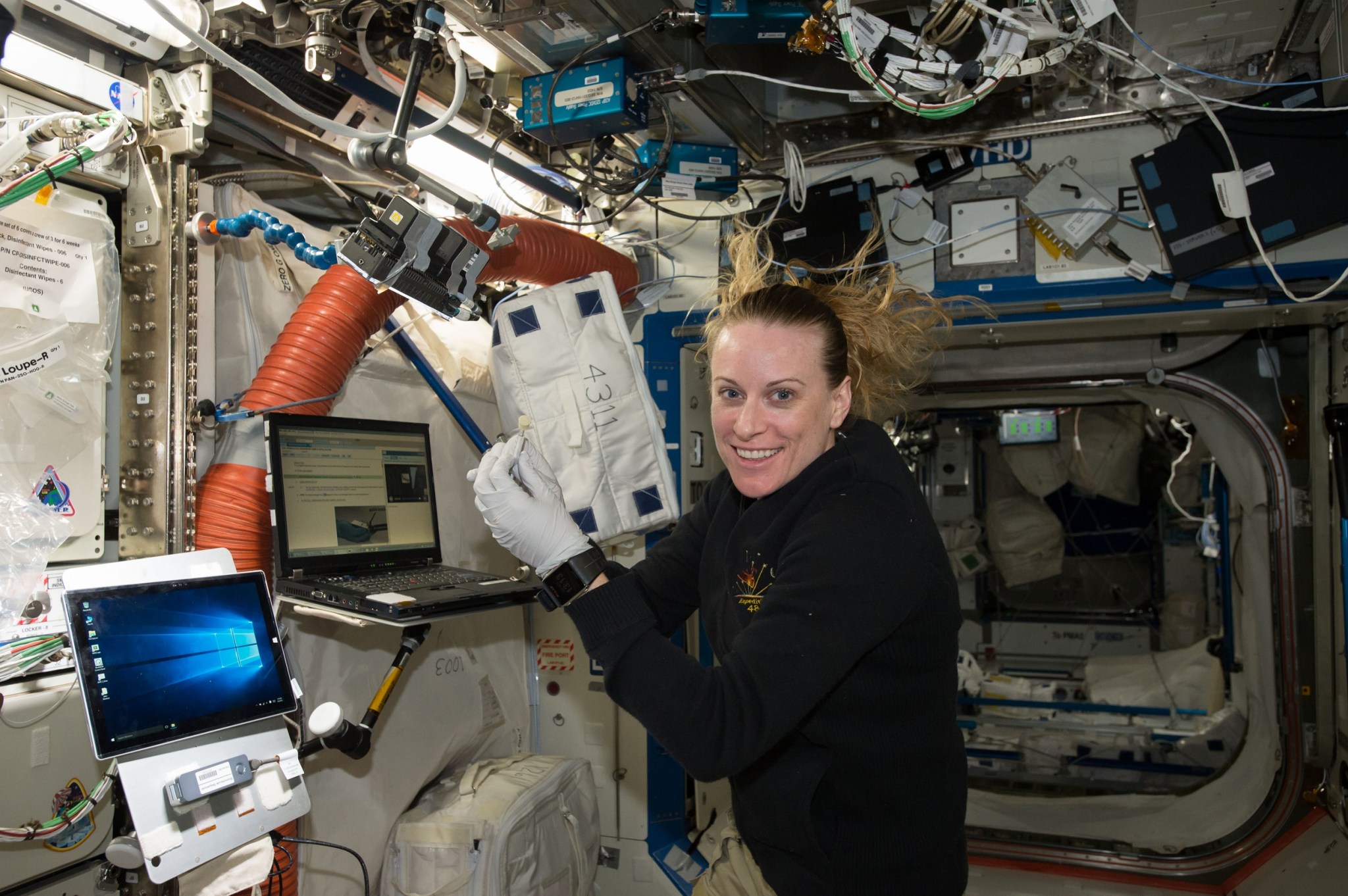Rubins wears a black sweatshirt as she holds a small experiment tube and smiles at the camera. There are two laptops in front of her and equipment and wiring above her.
