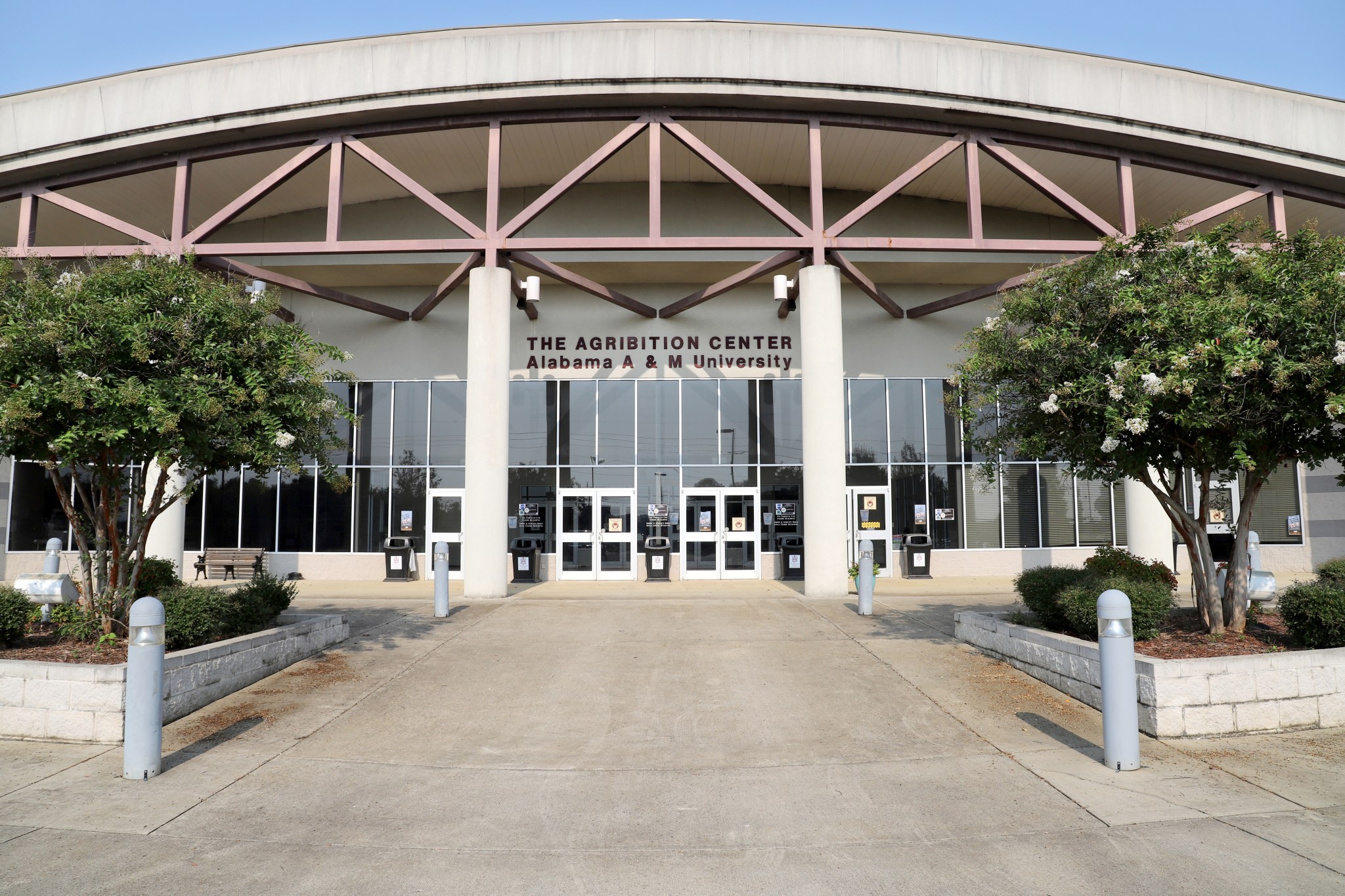 An external image of the Alabama A&M University Agribition Center from the front facade. The Center is a cream-colored stone building with a curved roof, floor-to-ceiling windows, and concrete steps that lead to a covered awning, framed by deep-red structural beams above. Shrubs and crepe myrtle trees frame the foreground and steps leading up to the building. Photo courtesy of AAMU Extension