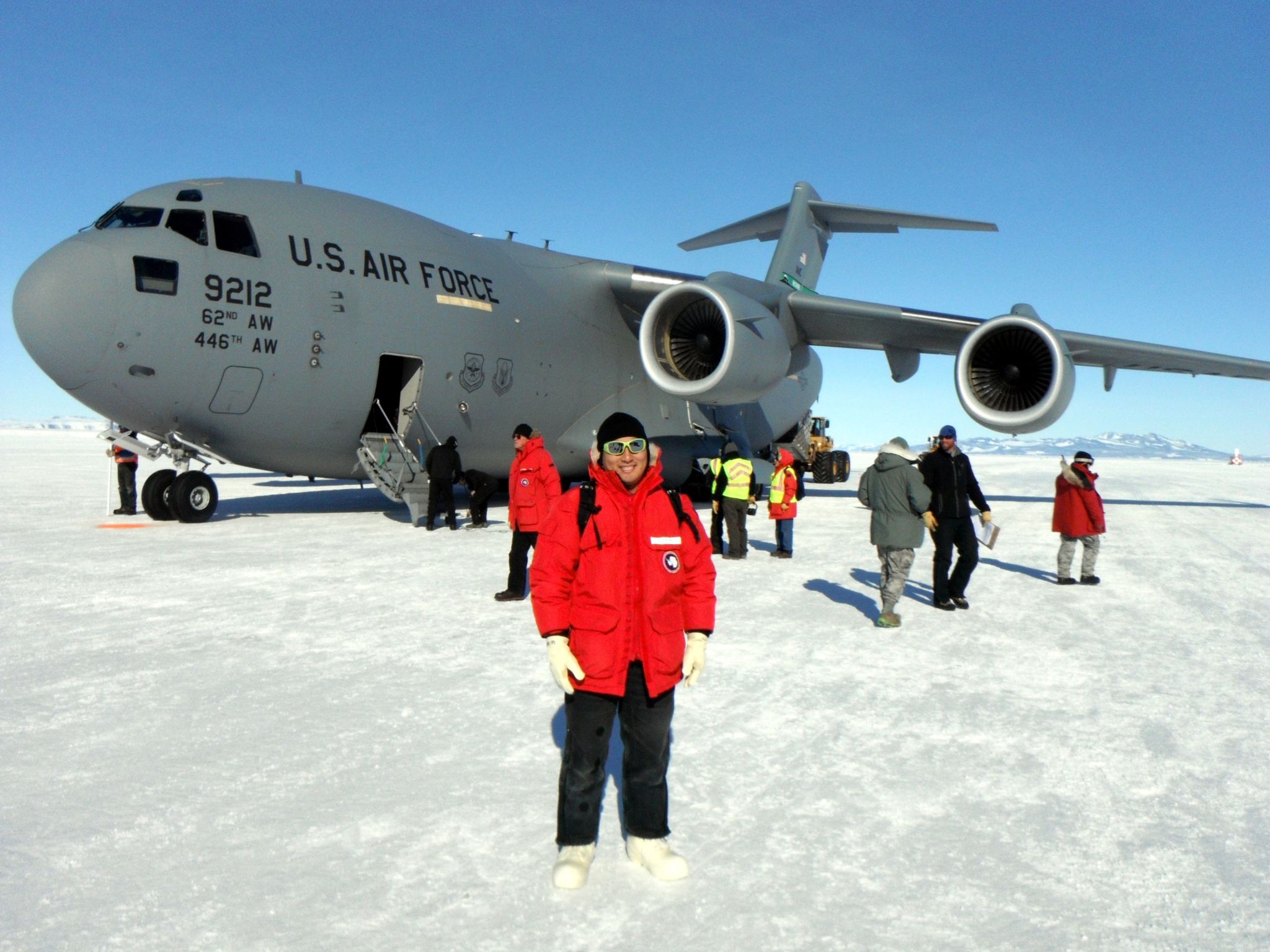 Kyle Helson stands in front of large grey C-17 airplane with "U.S. Airforce" in large black letters on the side. Kyle and the plane are on a snow and ice-covered ground. He wears a red coat with black pants. There are seven people working in the background. 