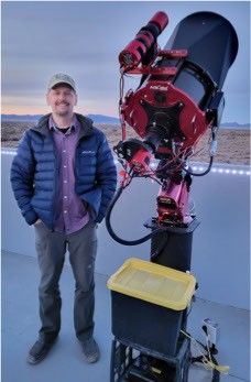 Man standing next to a large red telescope on an outdoor observatory platform at sunset.