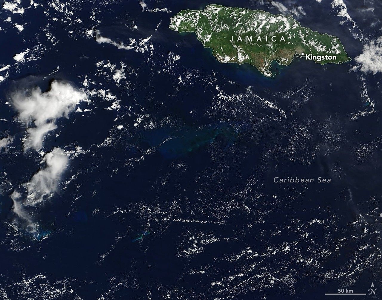 A satellite image shows a portion of the dark blue Caribbean Sea near Jamaica. A submerged carbonate platform appears as a slightly brighter blue area of water in the center. The mostly green island of Jamaica is in the upper right, and scattered clouds are present throughout.
