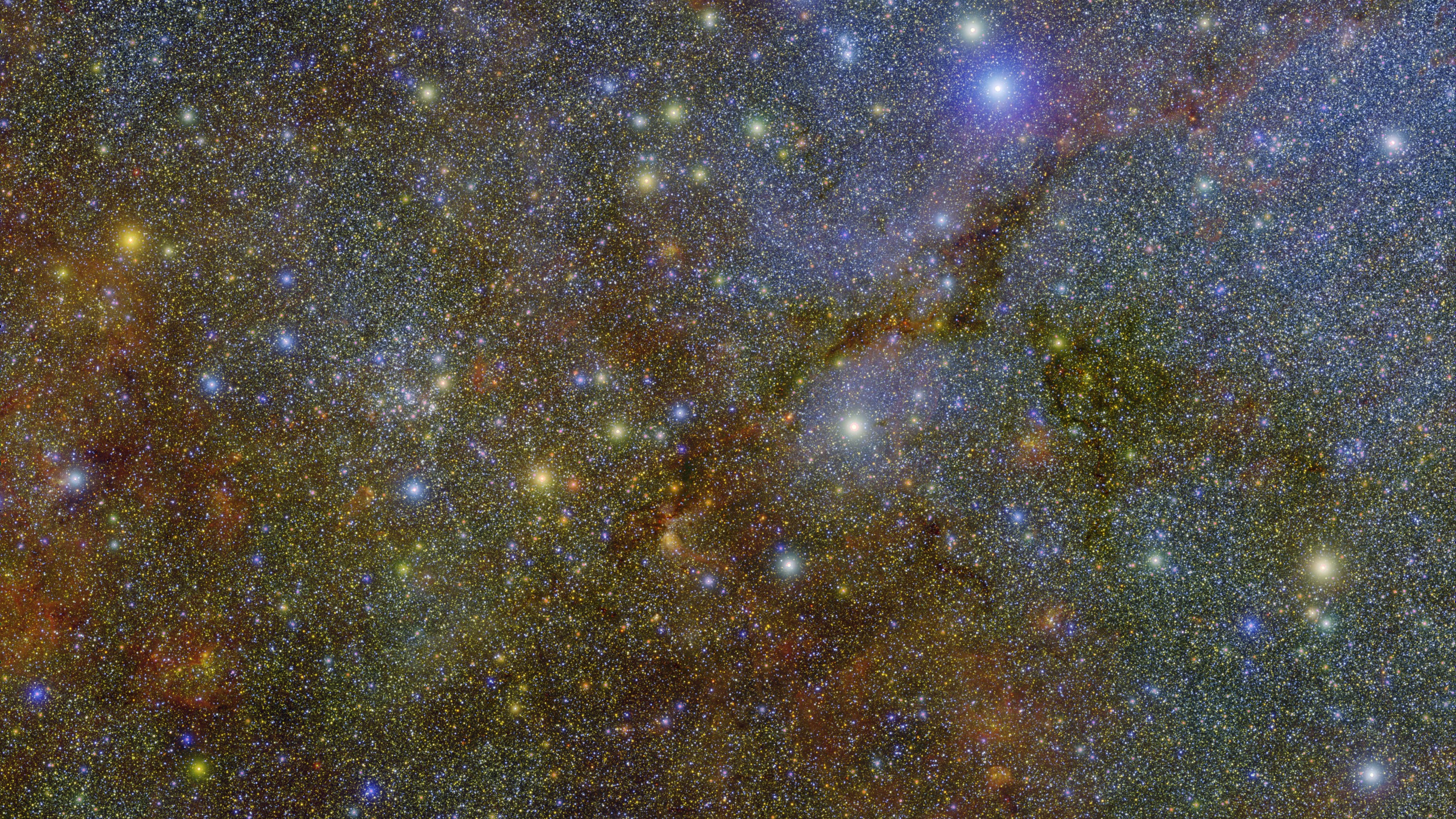 A dense tapestry of countless colorful stars fills the frame, punctuated by larger, brilliant points of light and faint, wispy veins of dark cosmic dust stretching across the deep space background.
