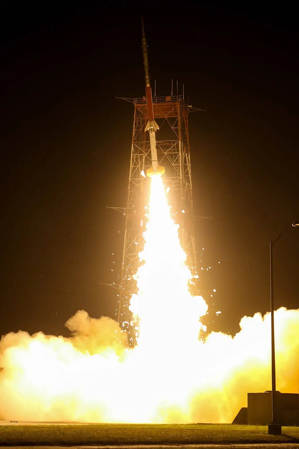 A rocket launches at night, surrounded by bright flames and smoke, with a tall supporting structure visible and the dark sky in the background.
